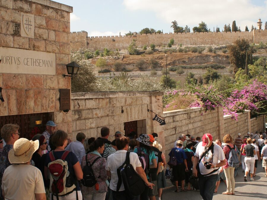 Older tourists in sun hats queue at the Hortus Gethsemani entrance to the Garden of Gethsemane, with bougainvillea on the wall and the Old City walls behind