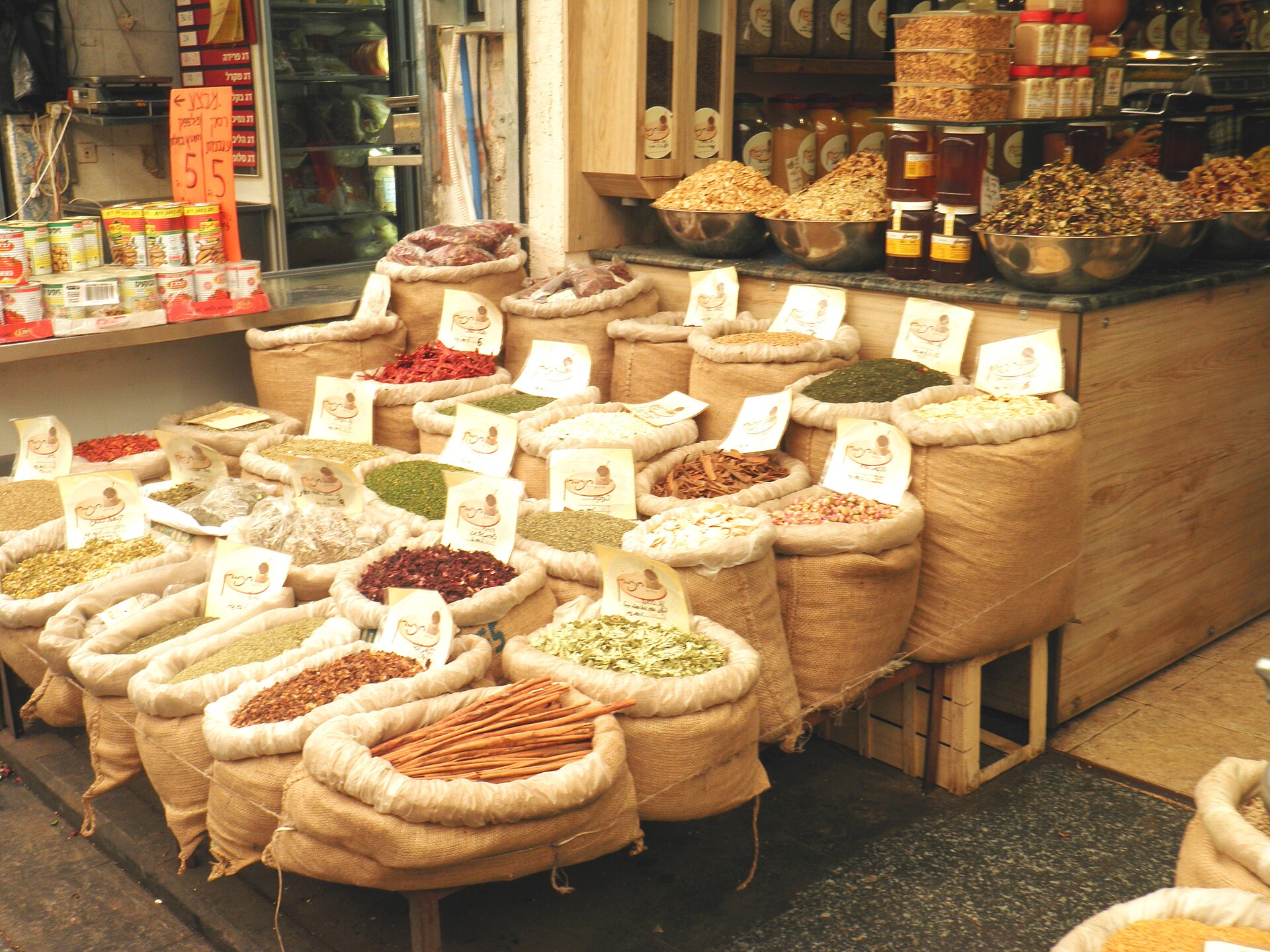 Open burlap sacks of cinnamon sticks, dried chillies, rosebuds, hibiscus and green herbs, each with a Hebrew label, at a spice shop in Mahane Yehuda Market