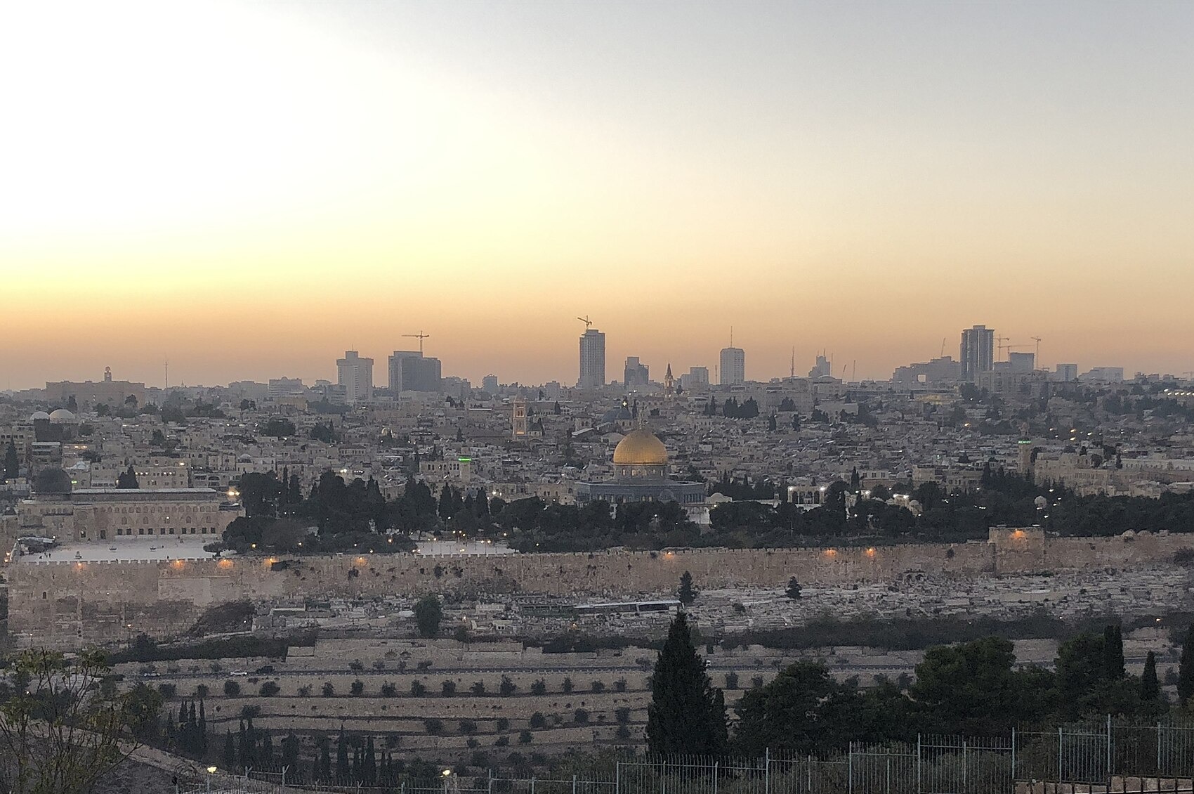 Orange sunset sky over Jerusalem's Old City, seen from the railed Mount of Olives lookout, with the golden Dome of the Rock, the eastern walls and cypresses below