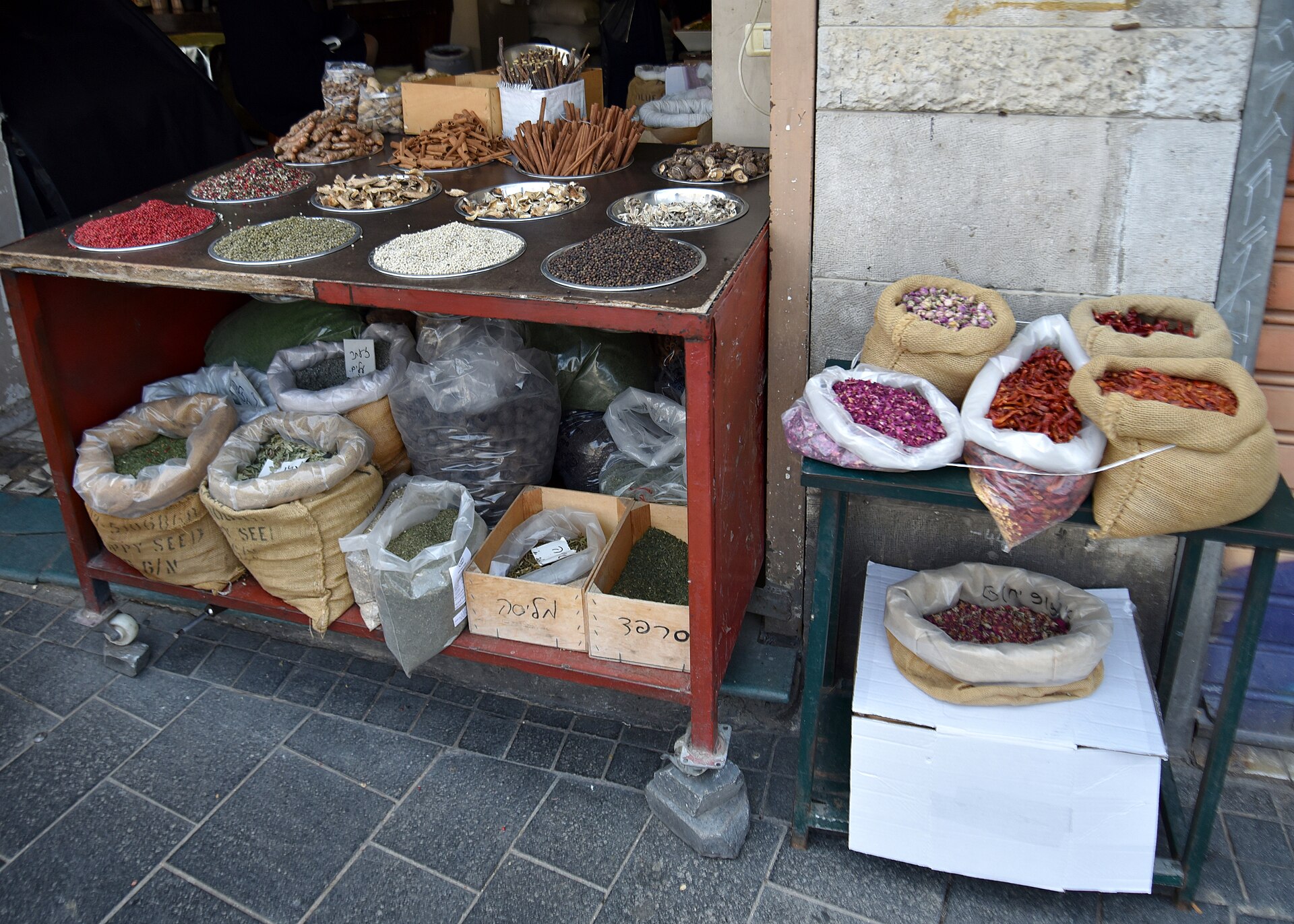 Pans of pink, green, white and black peppercorns, cinnamon bark and dried mushrooms above sacks of herbs and rosebuds at a Mahane Yehuda Market spice stall