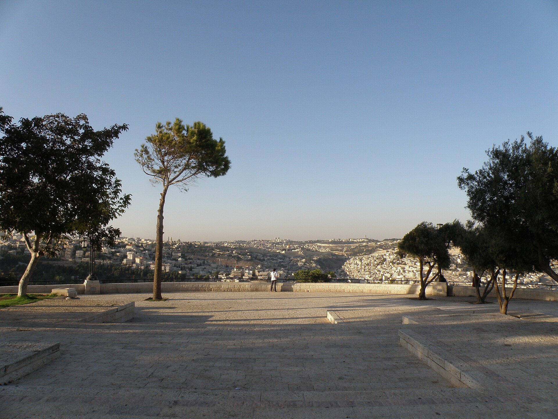 The paved stone terrace of the Haas Promenade in late-afternoon light, with a lone visitor at the railing looking north across the valley to the Old City