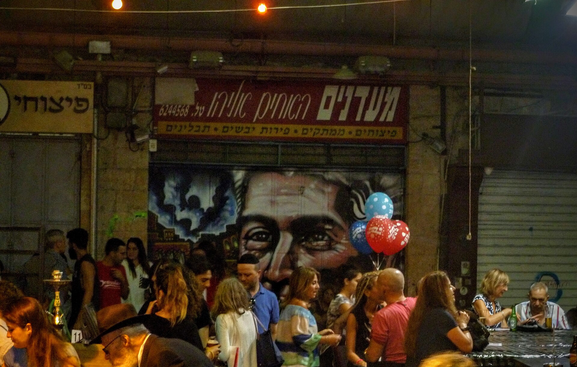 People drink and talk at tables after dark in front of a closed Mahane Yehuda stall whose shutter is painted with a large portrait, beneath its Hebrew shop sign