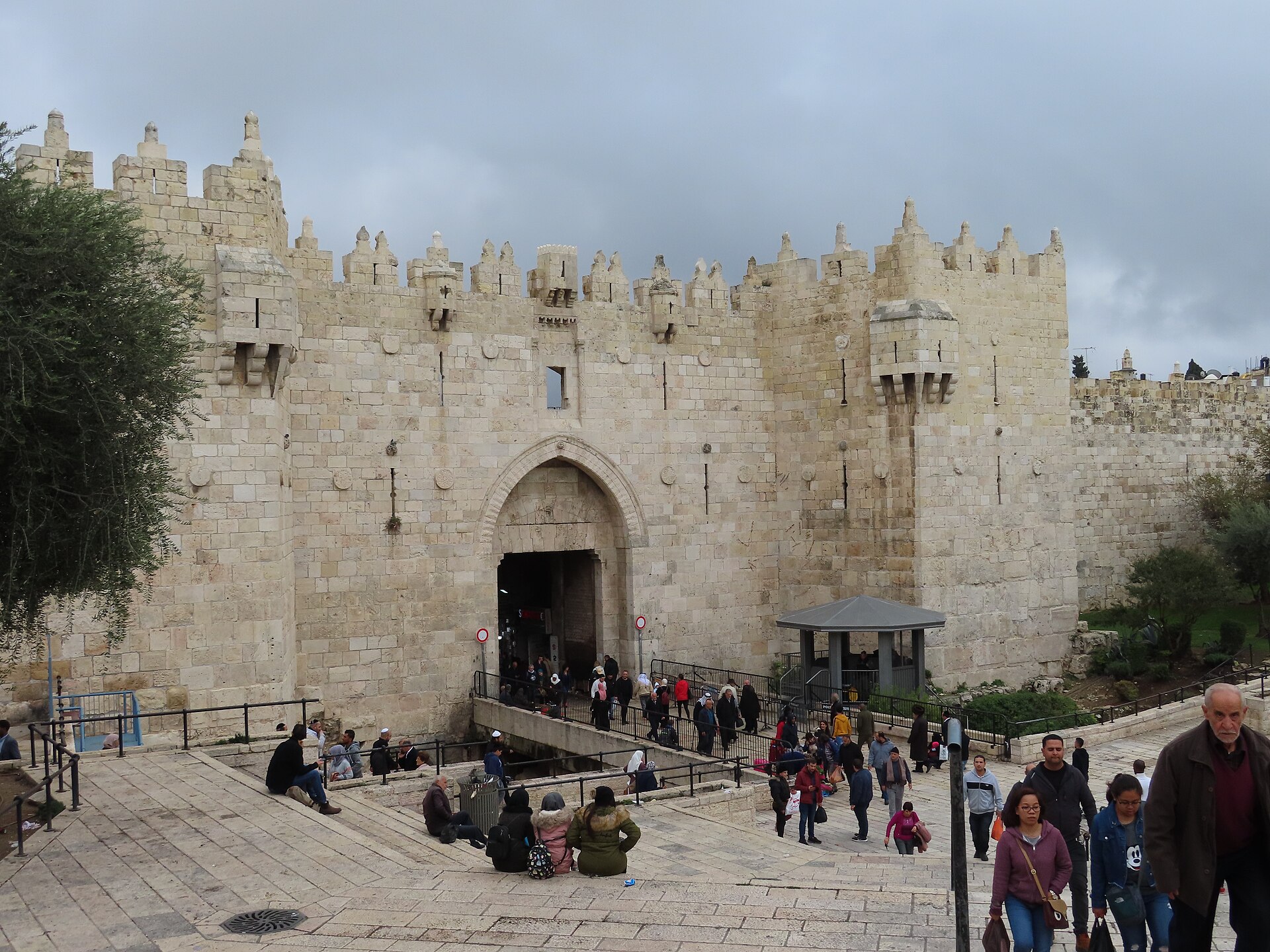 People sitting on the stone steps of the amphitheatre-like plaza facing Damascus Gate, with pedestrians crossing the bridge into the crenellated Ottoman gate