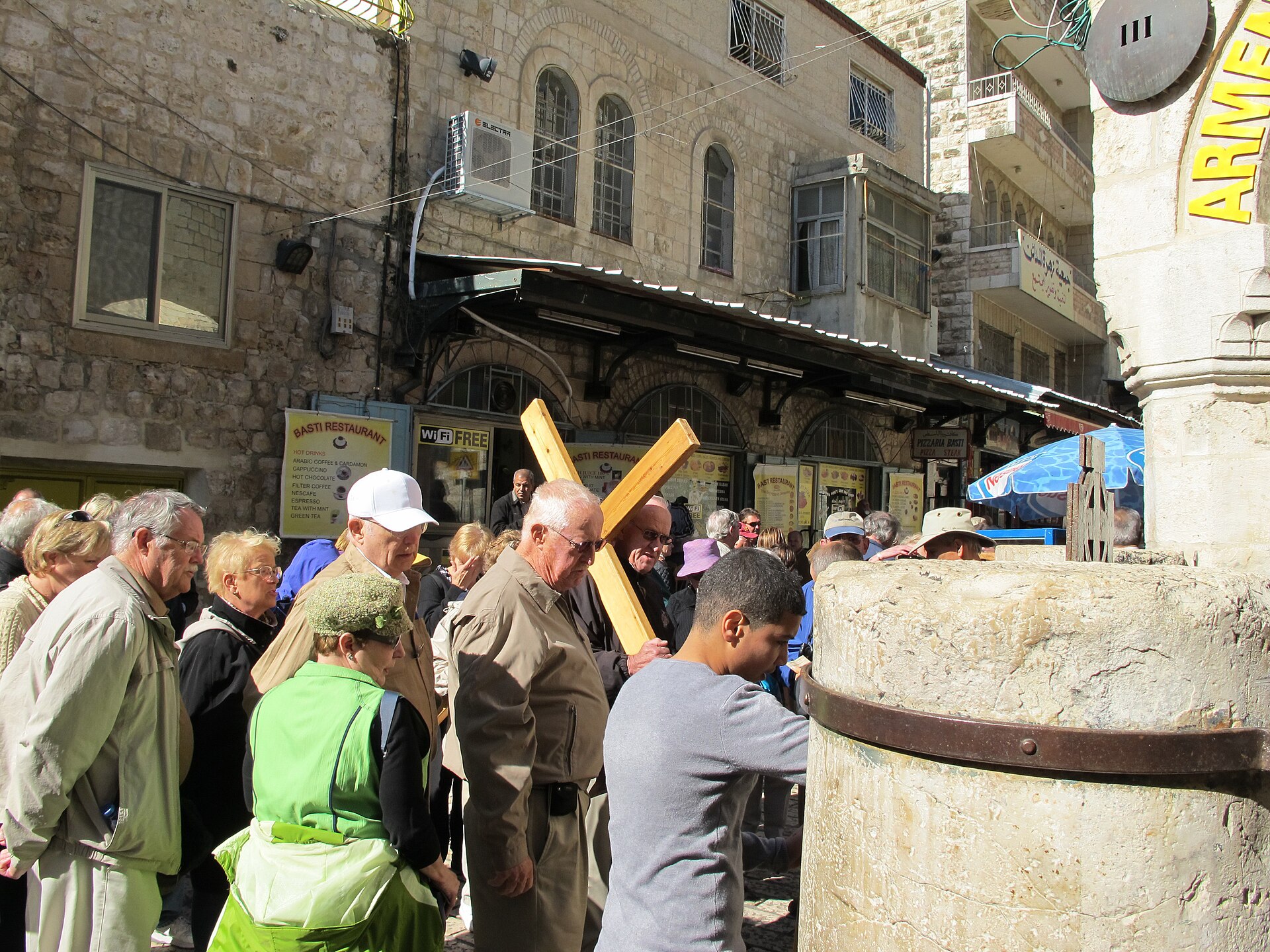 Pilgrim group, one man shouldering a light wooden cross, pausing at the Third Station of the Via Dolorosa beneath a round metal marker bearing the numeral III