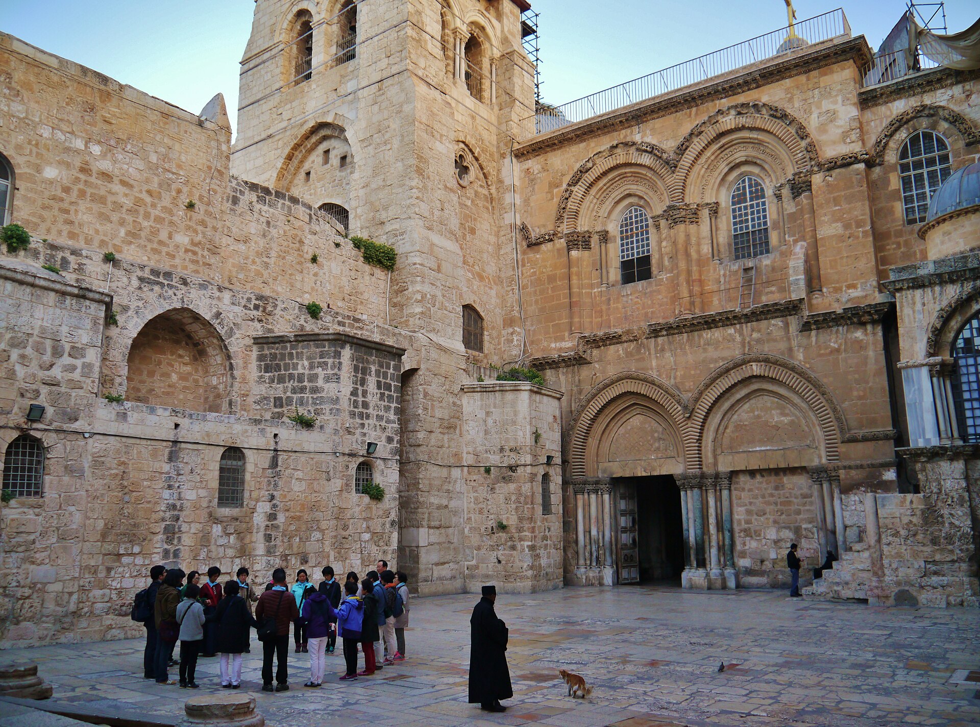 Pilgrim group standing in a circle in the quiet Holy Sepulchre parvis, below the bell tower and twin-arched Crusader entrance, with an Orthodox cleric nearby