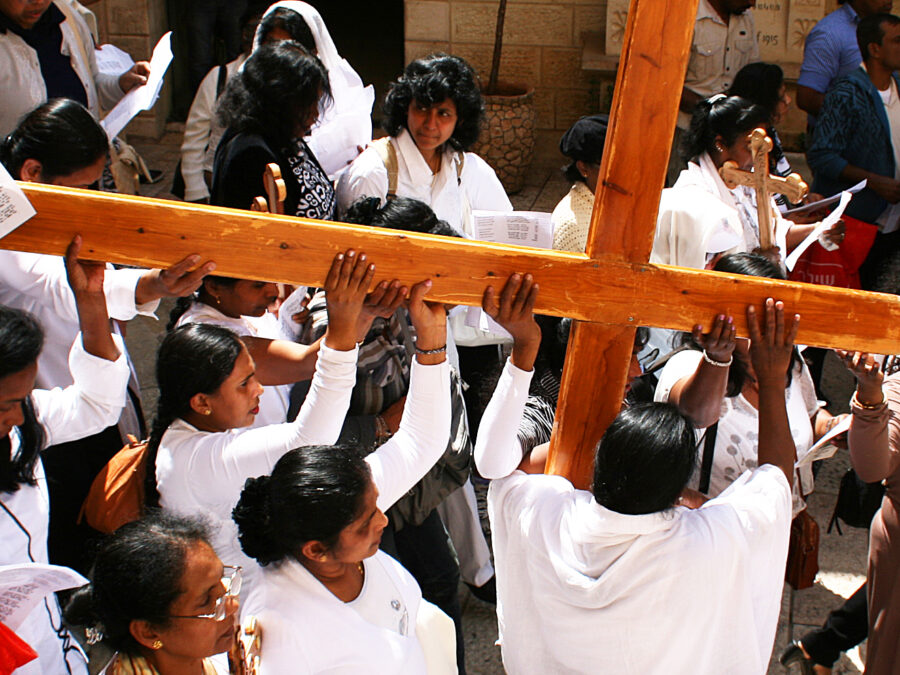 Pilgrims dressed in white lifting a large wooden cross above their heads while walking the Via Dolorosa in Jerusalem's Old City on Good Friday