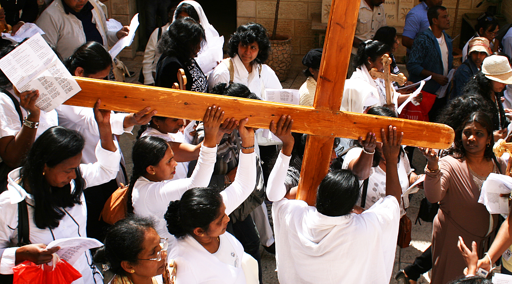 Pilgrims dressed in white lifting a large wooden cross above their heads while walking the Via Dolorosa in Jerusalem's Old City on Good Friday