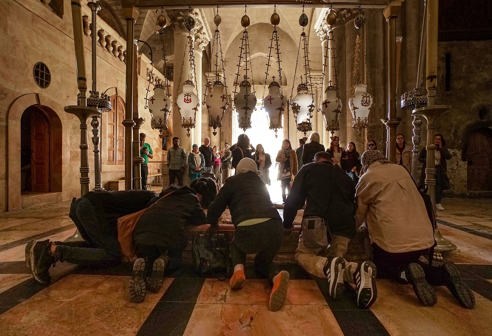 Pilgrims kneeling to touch the Stone of Anointing beneath a row of hanging lamps, with the bright entrance doorway of the Church of the Holy Sepulchre behind