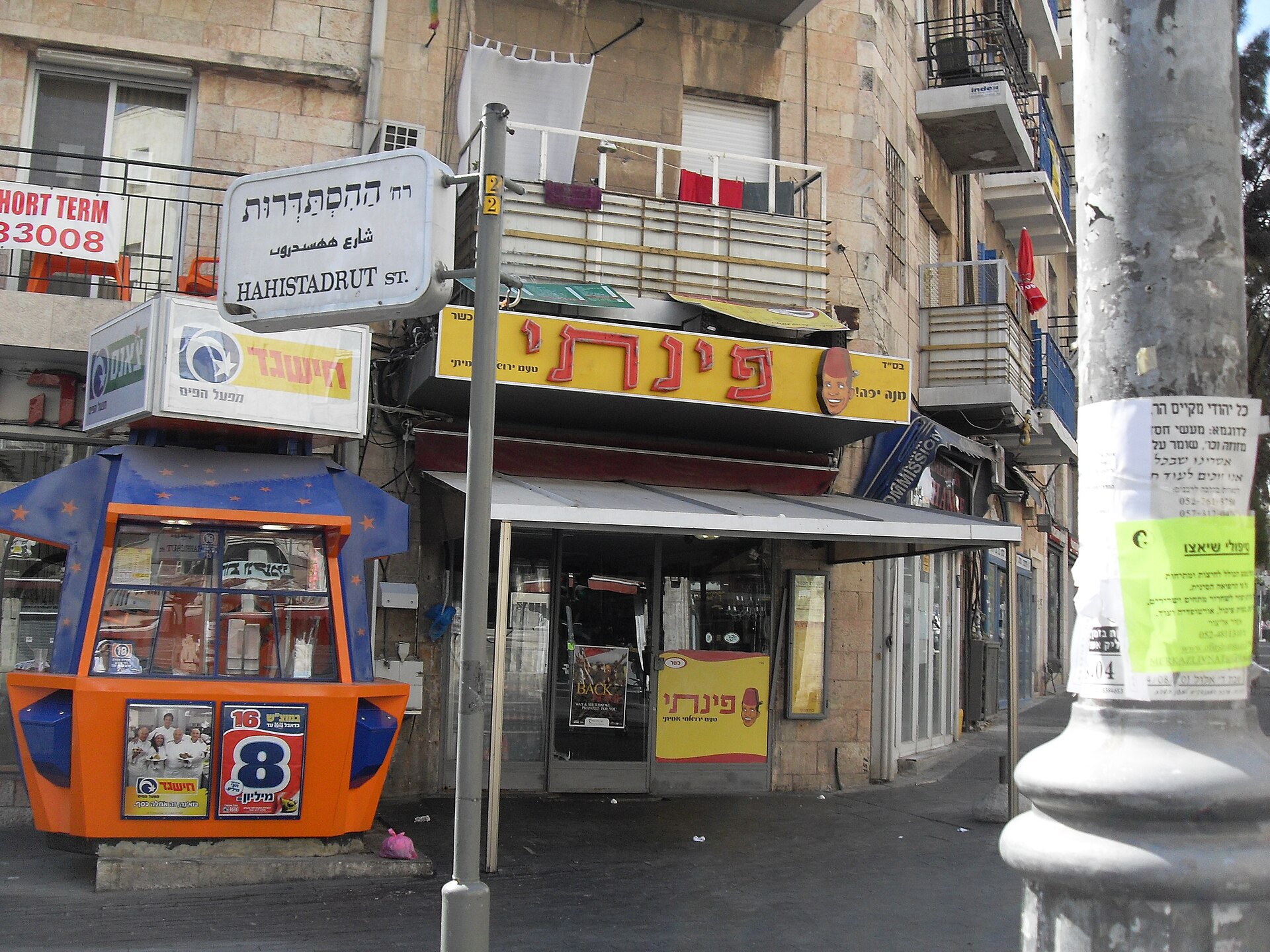 Pinati hummus restaurant's yellow Hebrew sign above its corner shopfront on King George Street, with an orange lottery kiosk and Hahistadrut Street sign in front