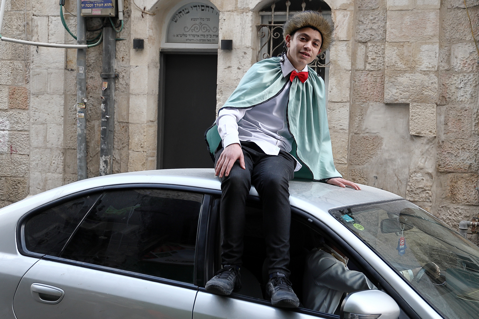 A young man in a fur shtreimel hat and pale green cape sitting on a car roof in a Jerusalem street during Purim