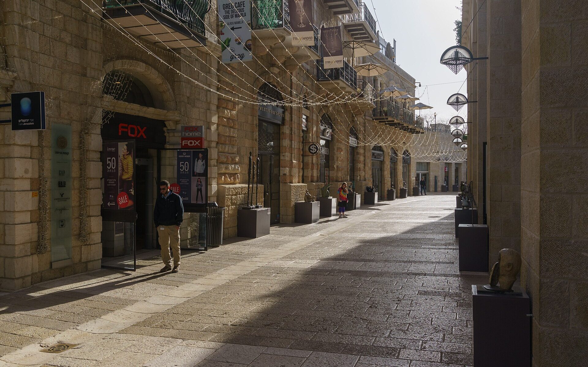 Quiet morning on Mamilla Avenue in Jerusalem, with restored stone buildings, iron balconies, shopfronts, strings of lights overhead and bronze sculptures on plinths along the paved street