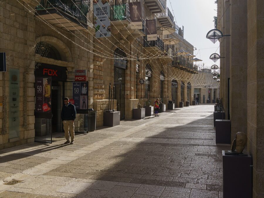 Quiet morning on Mamilla Avenue in Jerusalem, with restored stone buildings, iron balconies, shopfronts, strings of lights overhead and bronze sculptures on plinths along the paved street
