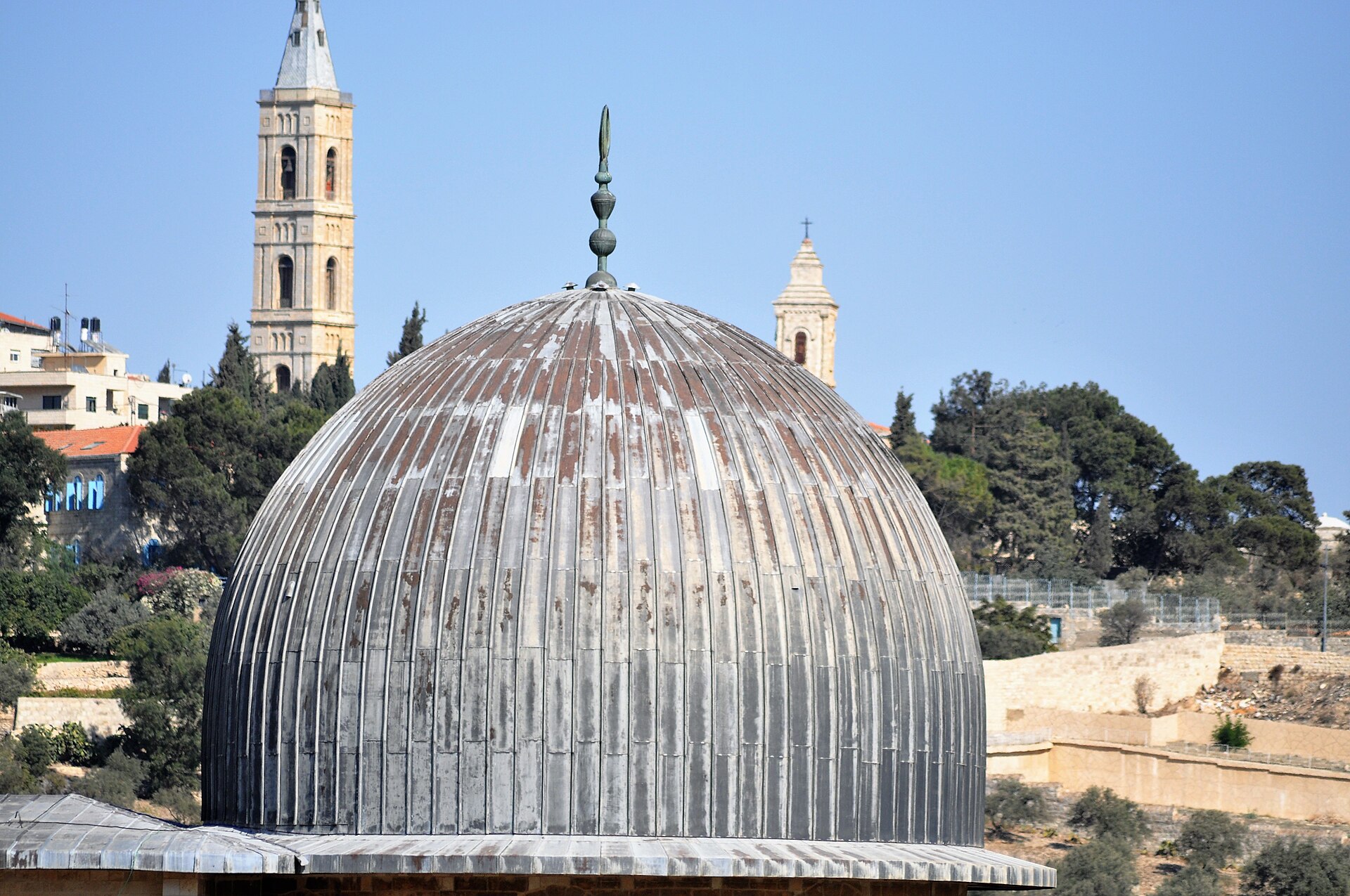 The ribbed grey lead dome of the Al-Aqsa Mosque, topped by a crescent finial, with church bell towers on the Mount of Olives behind