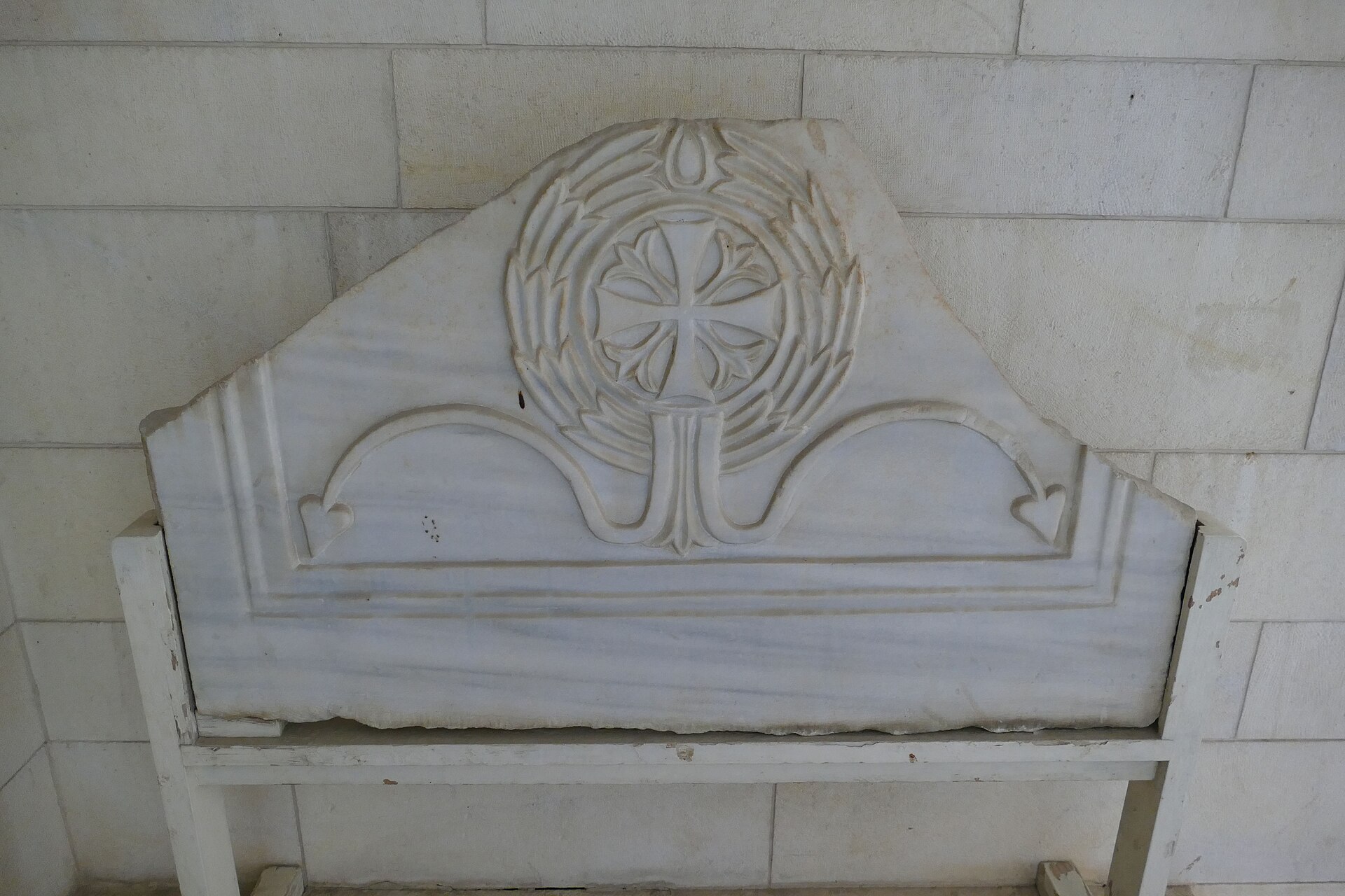 A carved white marble panel with a cross inside a wreath and scrolling tendrils, propped on a wooden stand against a stone wall at the Rockefeller Museum in Jerusalem