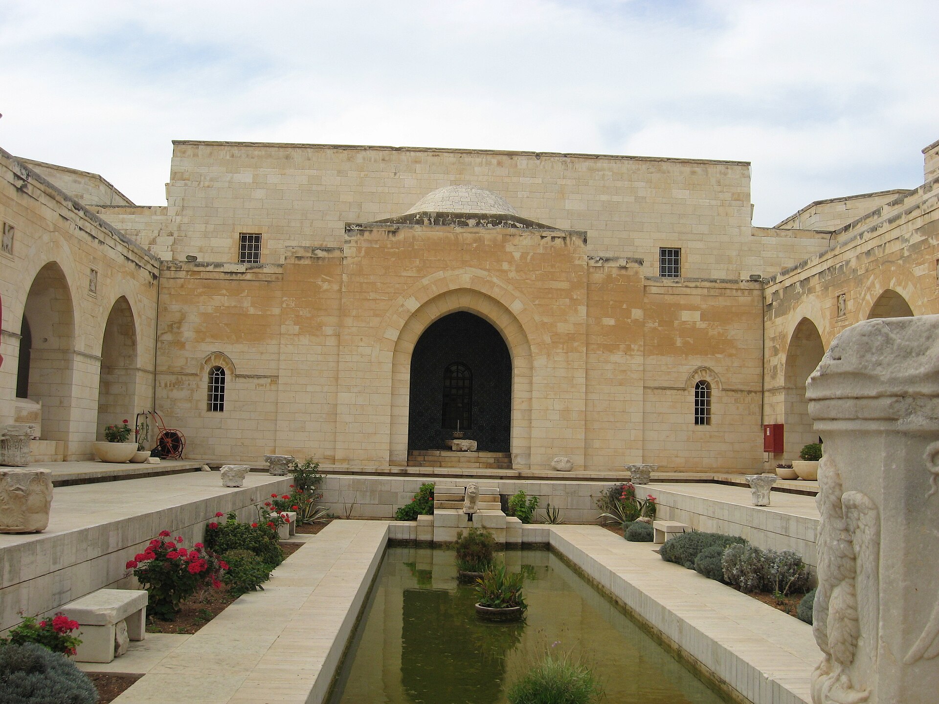The inner cloister of the Rockefeller Museum in Jerusalem, with a long rectangular pool, flowering plants, ancient column capitals set along the edges, and a tiled arched doorway at the far end