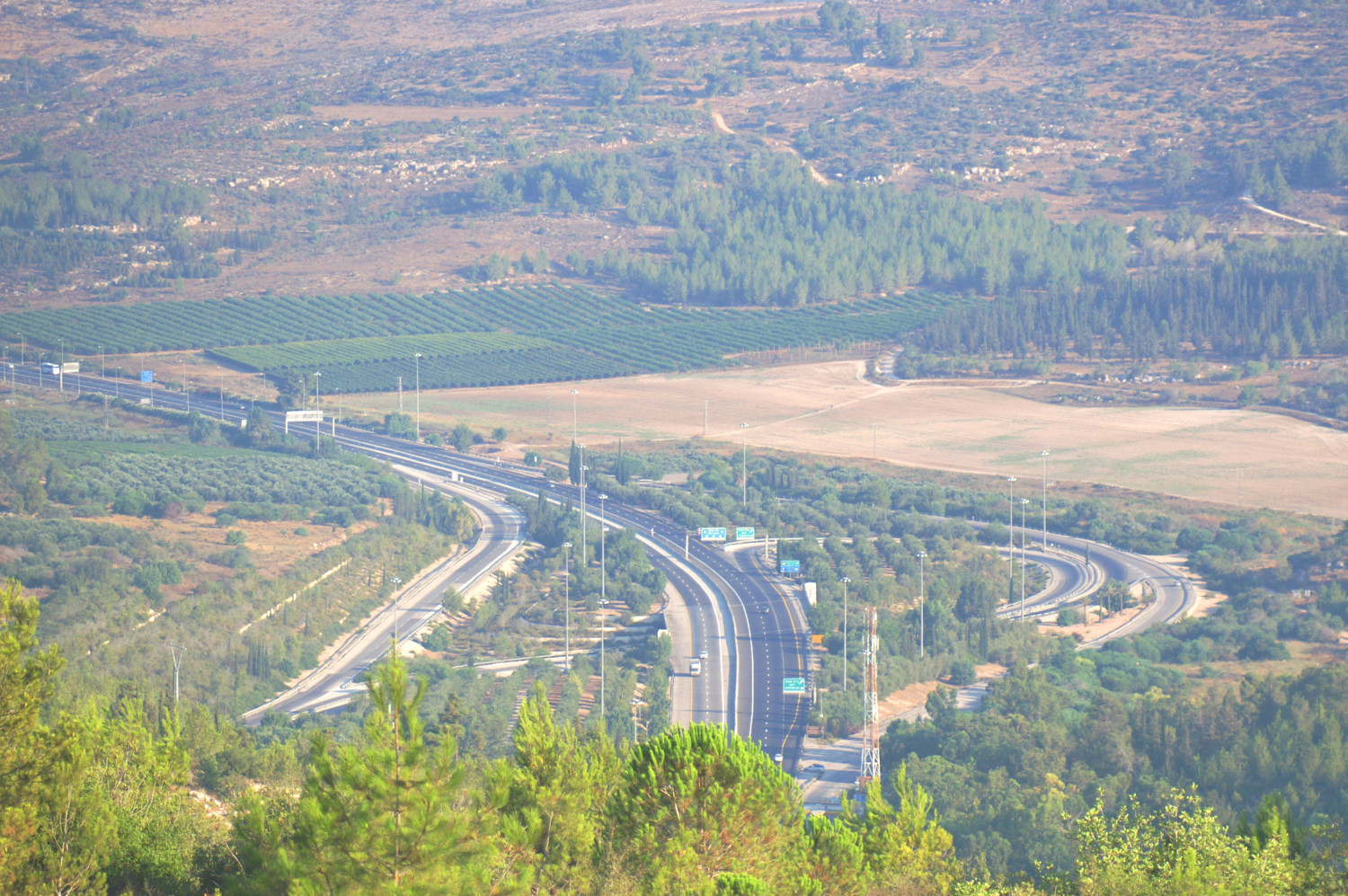 Route 1 motorway curving through the Sha'ar HaGai interchange below forested hills, with orchards and fields at the foot of the Judean Hills