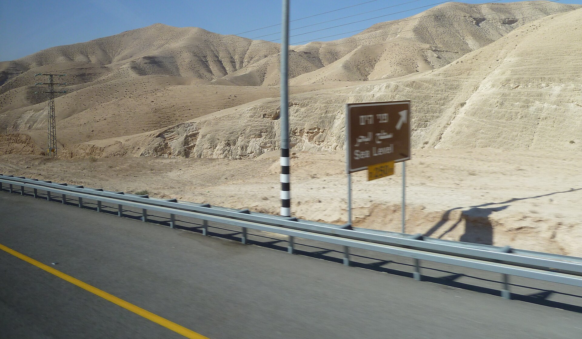 A highway winding through bare, sunlit hills in the Judean Desert, with a brown road sign beside the crash barrier