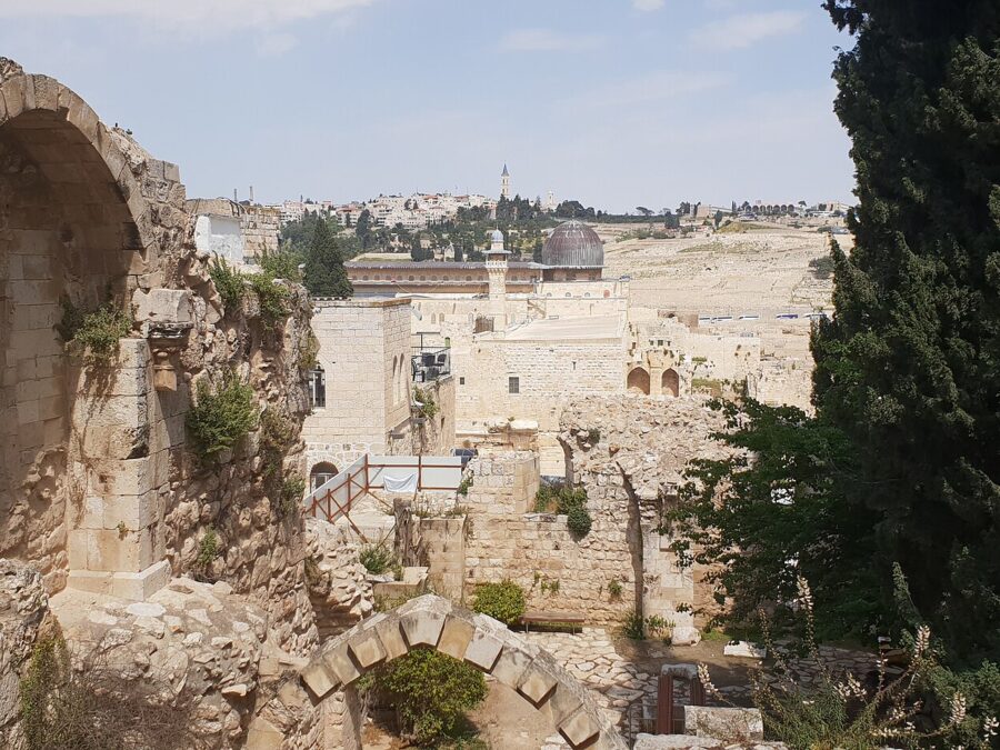 Ruined apse and pointed arch of the Crusader church of St Mary of the Germans, with the grey dome of al-Aqsa Mosque and the Mount of Olives beyond