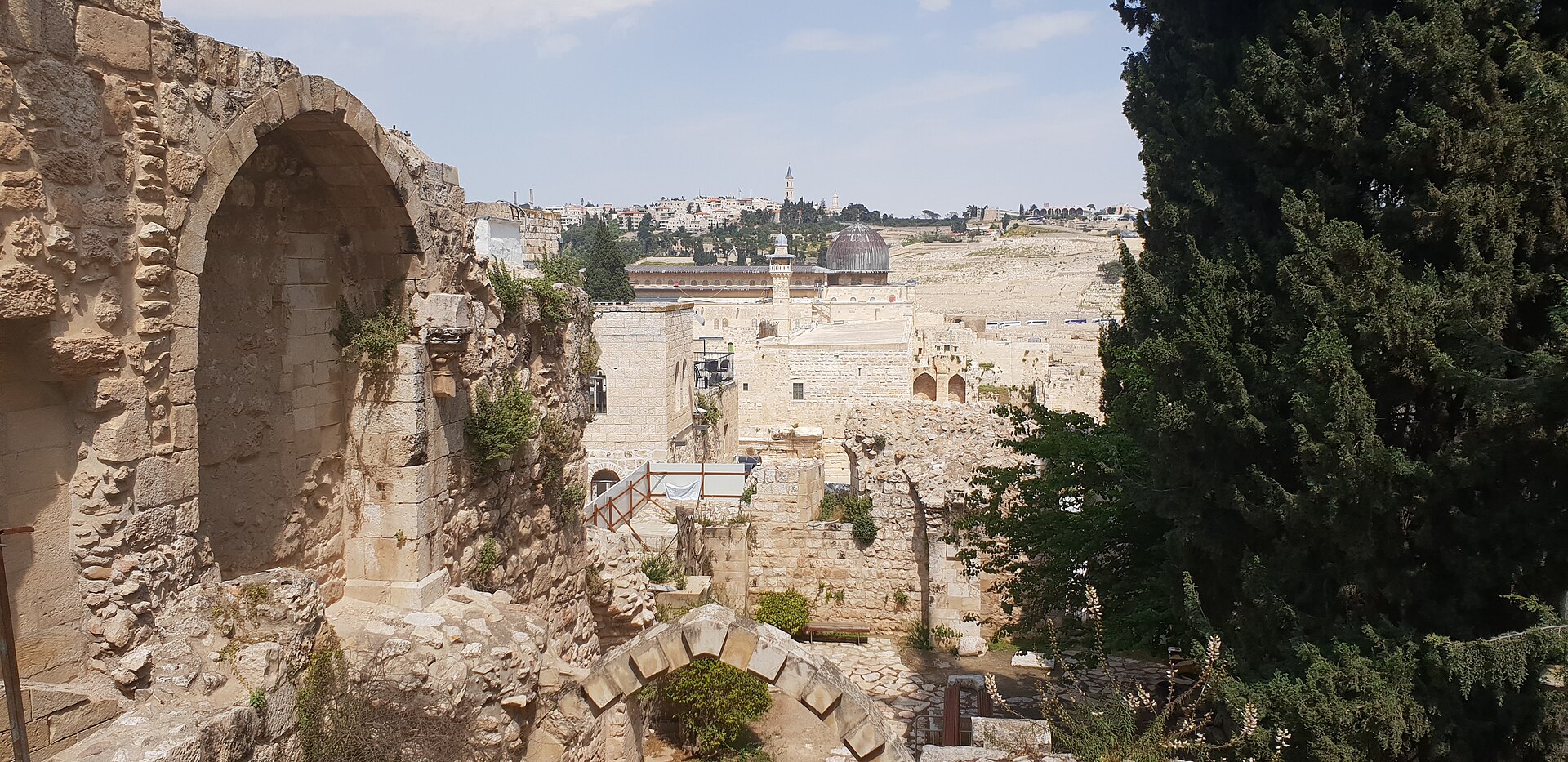 Ruined apse and pointed arch of the Crusader church of St Mary of the Germans, with the grey dome of al-Aqsa Mosque and the Mount of Olives beyond