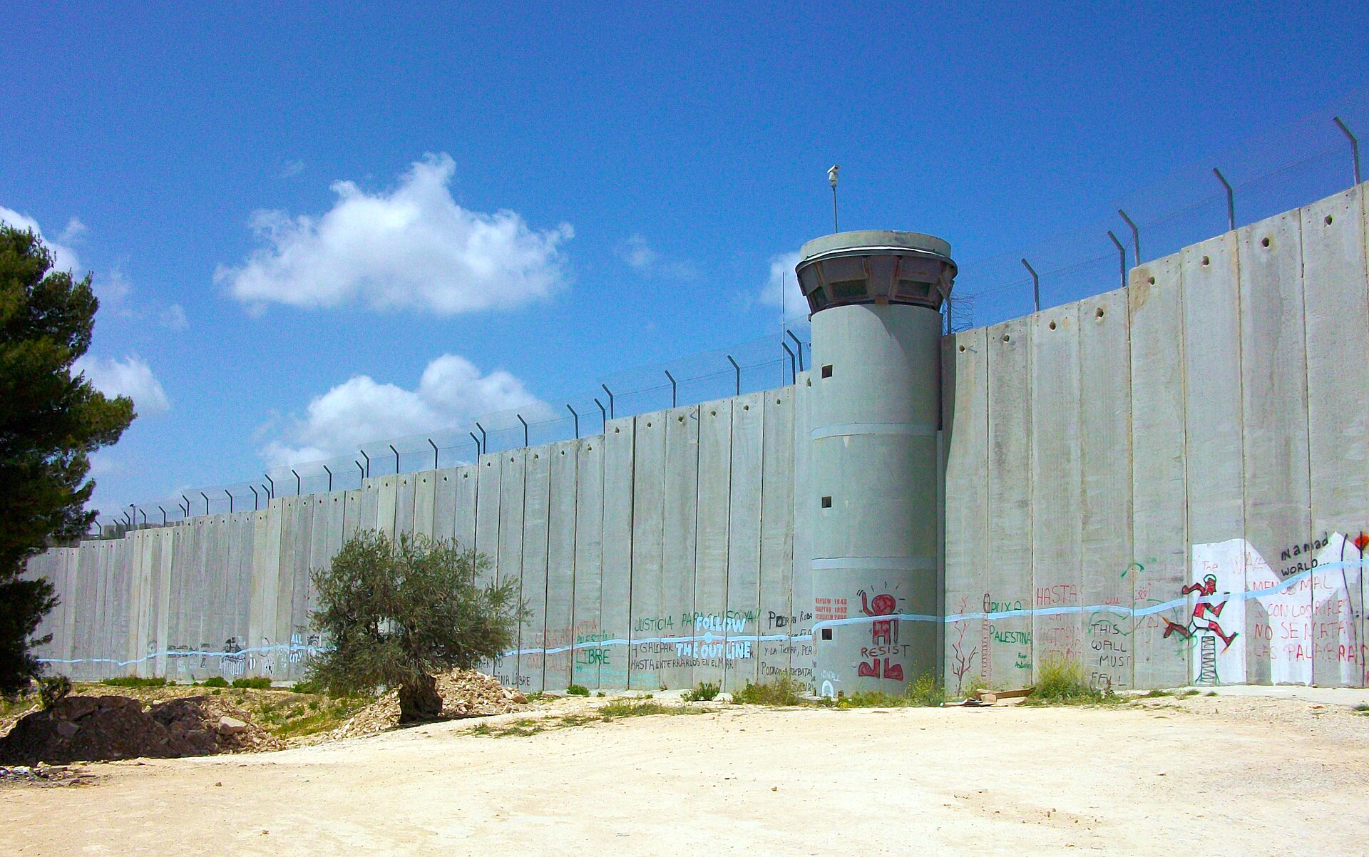 A tall grey concrete section of the separation barrier in Bethlehem with a round watchtower, barbed wire on top and painted slogans and murals along its base