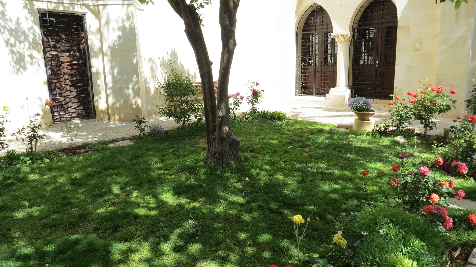 Shaded lawn with a tree and red and pink roses in the Christ Church compound near Jaffa Gate, in front of pale stone walls with arched iron-grilled doors
