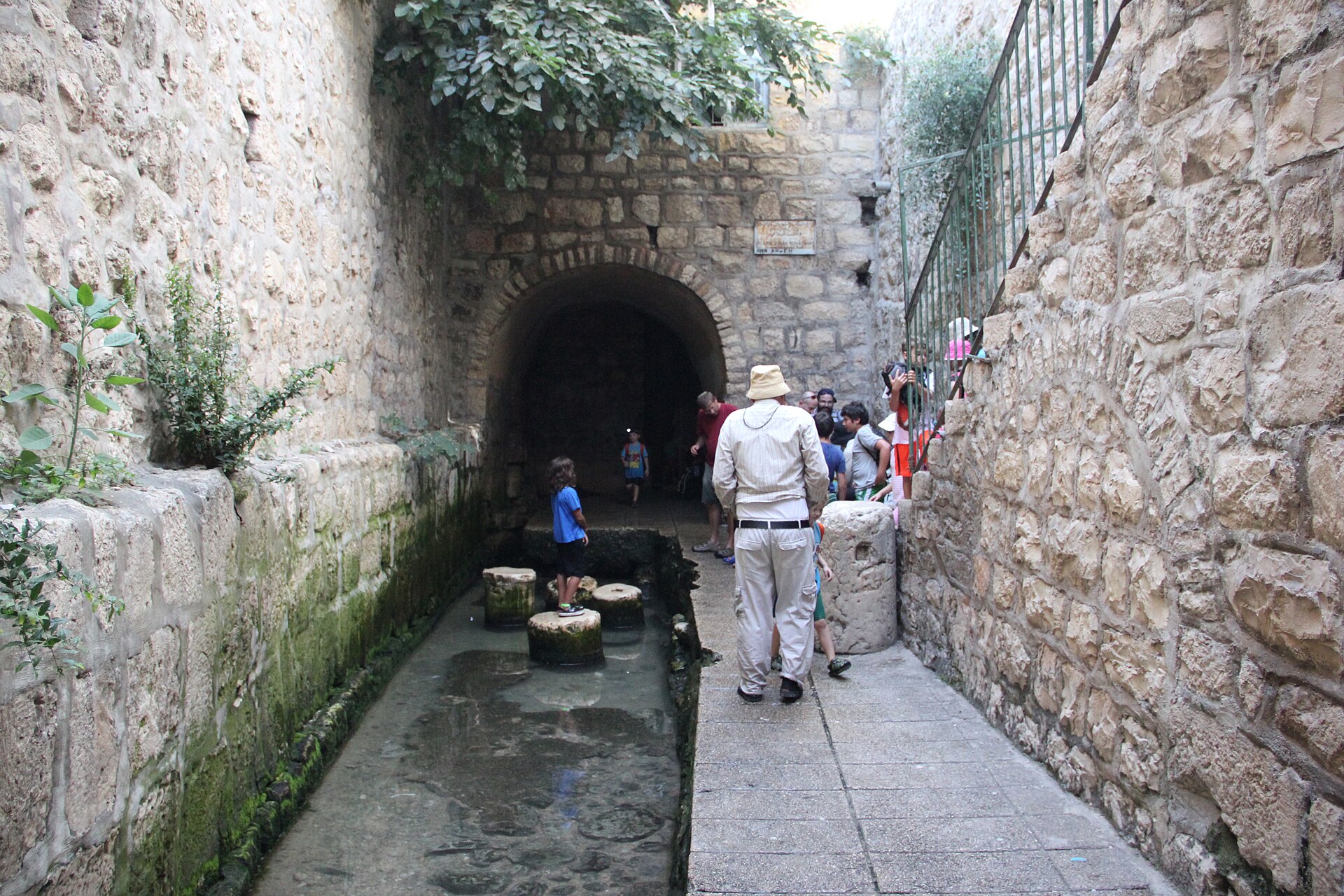 Shallow pool between high stone walls at the exit of Hezekiah's Tunnel, with old column drums in the water and visitors walking out from the arched tunnel