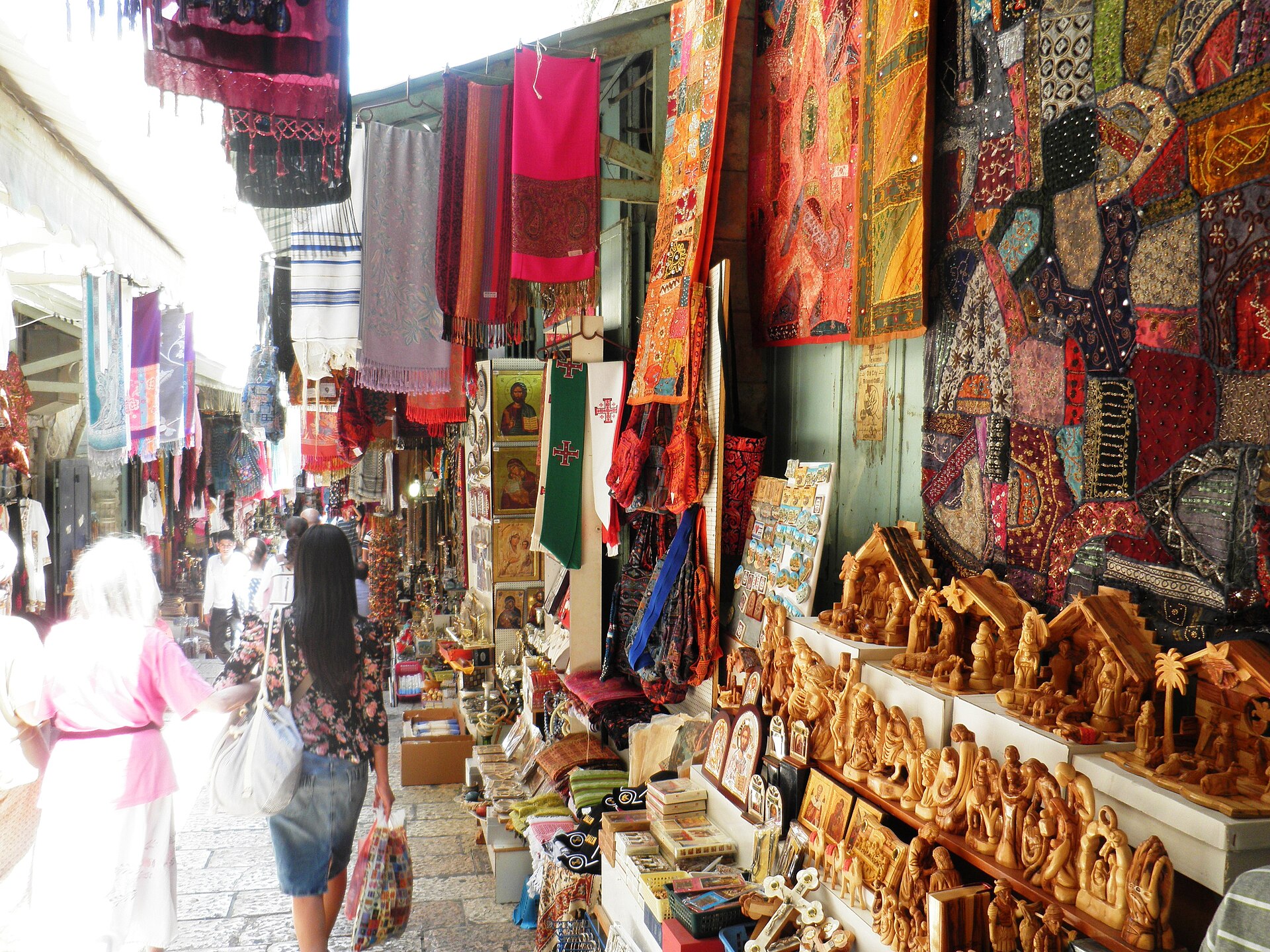 Shelves of carved olive-wood nativity scenes and figures at a David Street souvenir stall in Jerusalem's Old City, under embroidered hangings, icons and prayer shawls