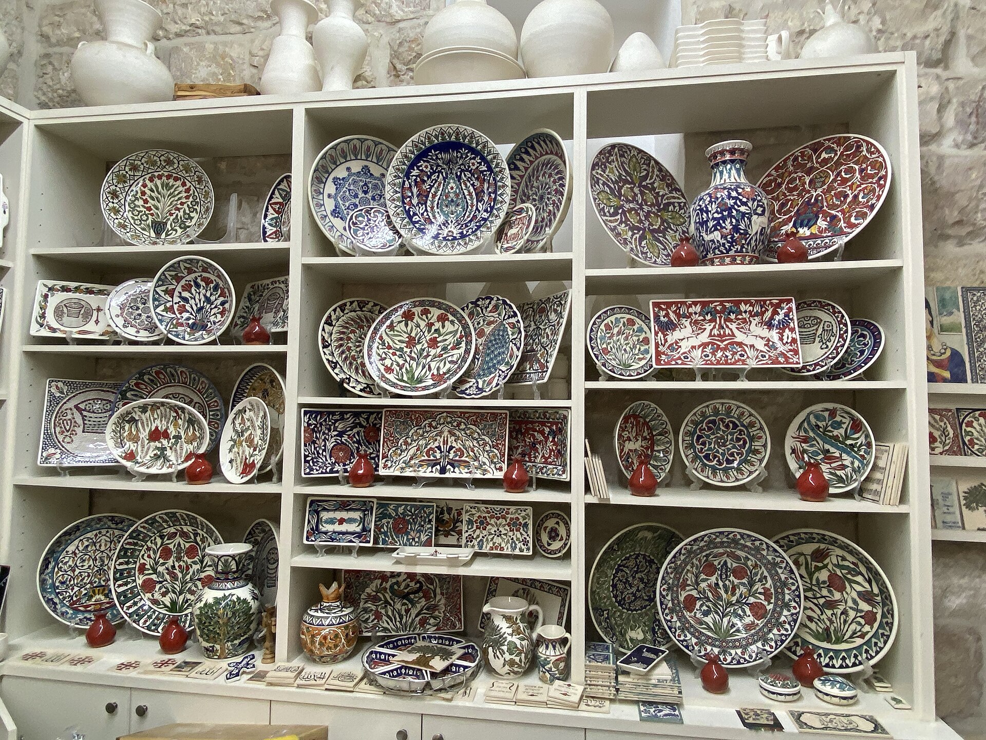Shelves of hand-painted Armenian ceramic plates, platters and vases with floral and bird designs and small red pomegranates in a stone-walled Jerusalem Old City workshop