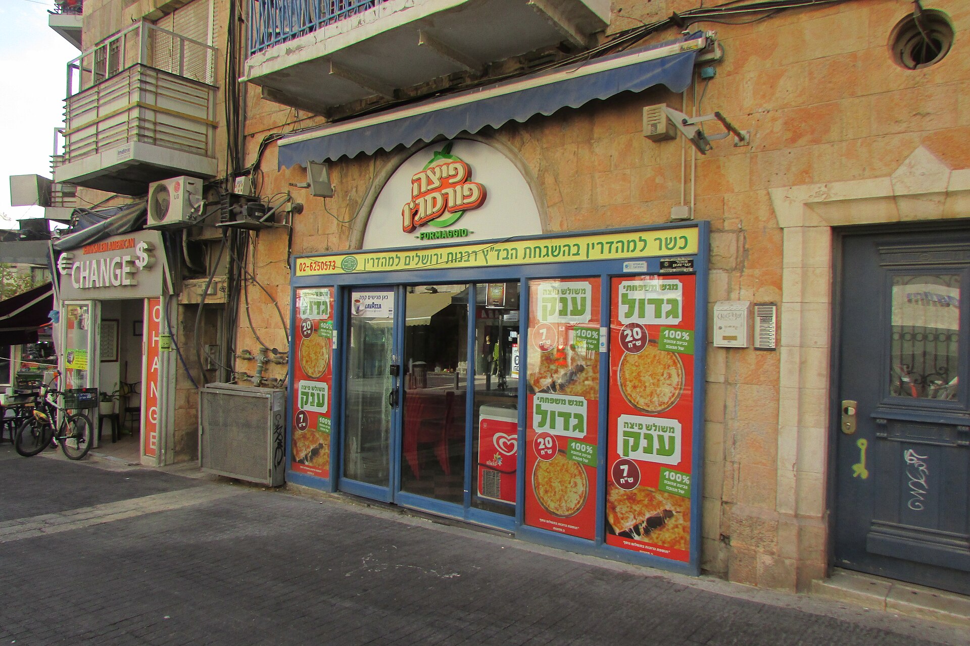 Shopfront of a kosher pizzeria on King George Street in Jerusalem, with Hebrew posters advertising family-size pizza trays and giant slices