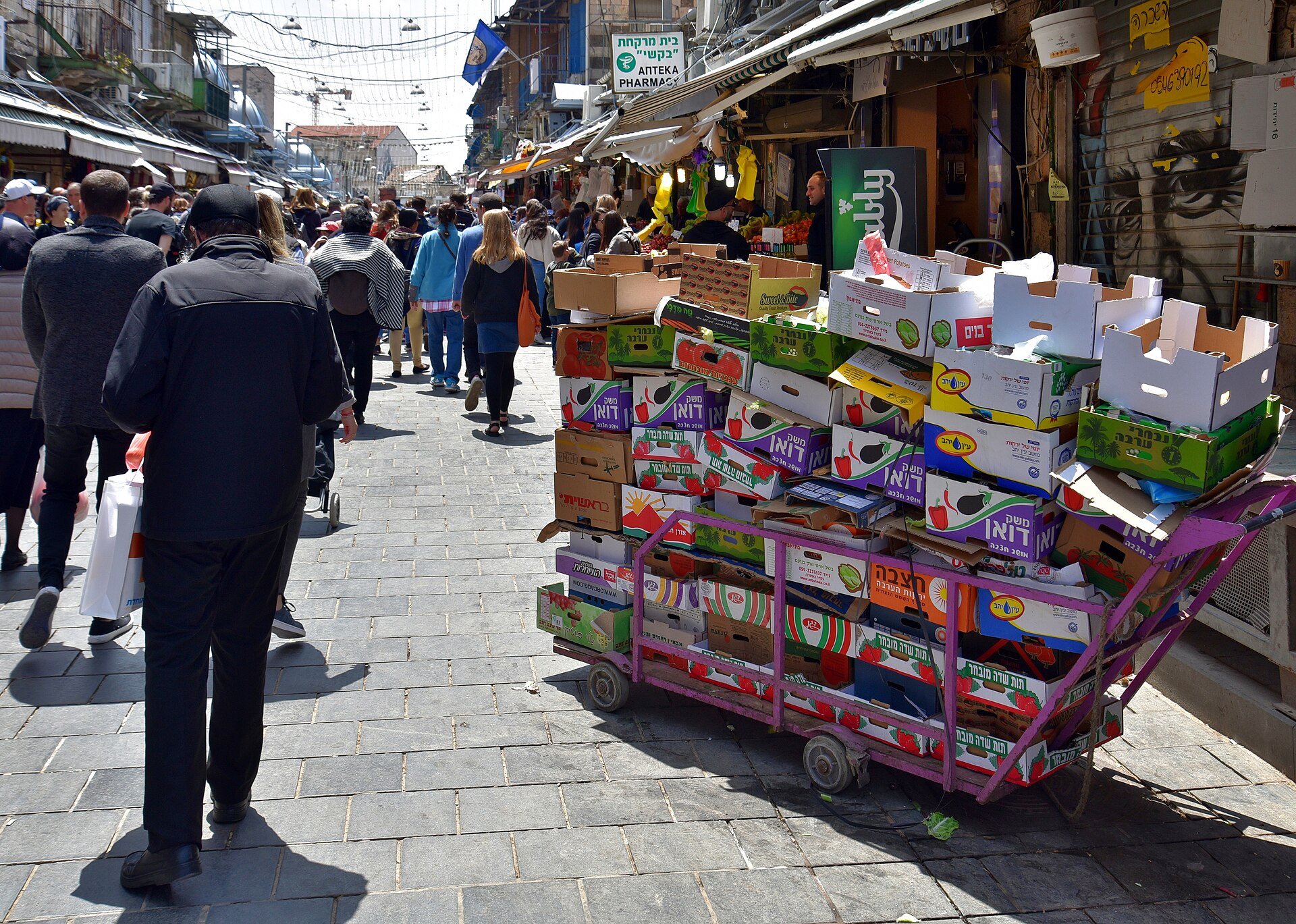 Shoppers crowd the open-air Mahane Yehuda Street past a hand-cart piled high with empty produce boxes, under shop awnings and overhead light strings