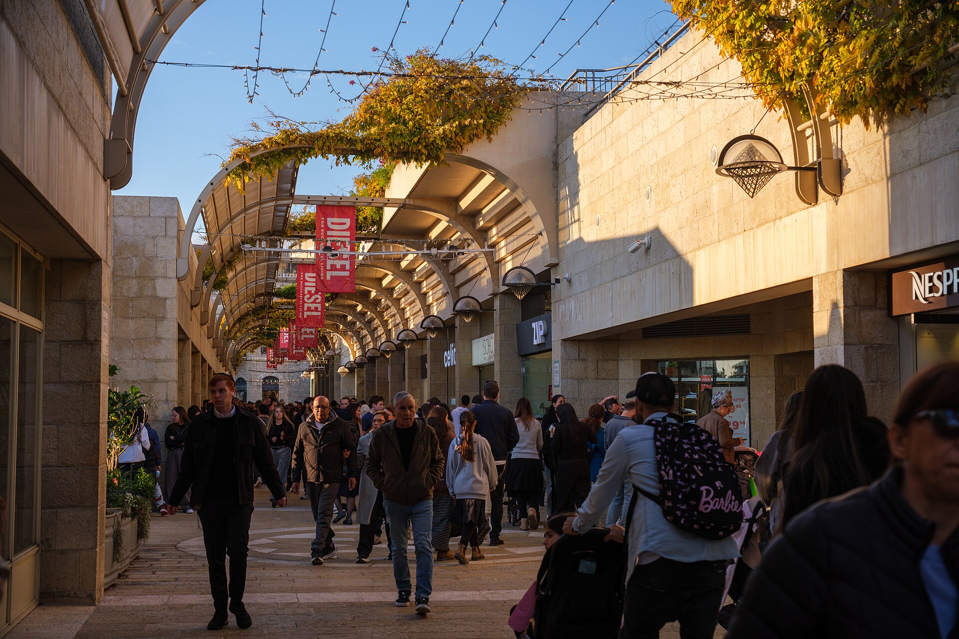 Shoppers and parents with pushchairs walking beneath the arched vine-covered canopy of Mamilla Avenue in Jerusalem, lined with Nespresso, Zip and Celio shops and Diesel banners