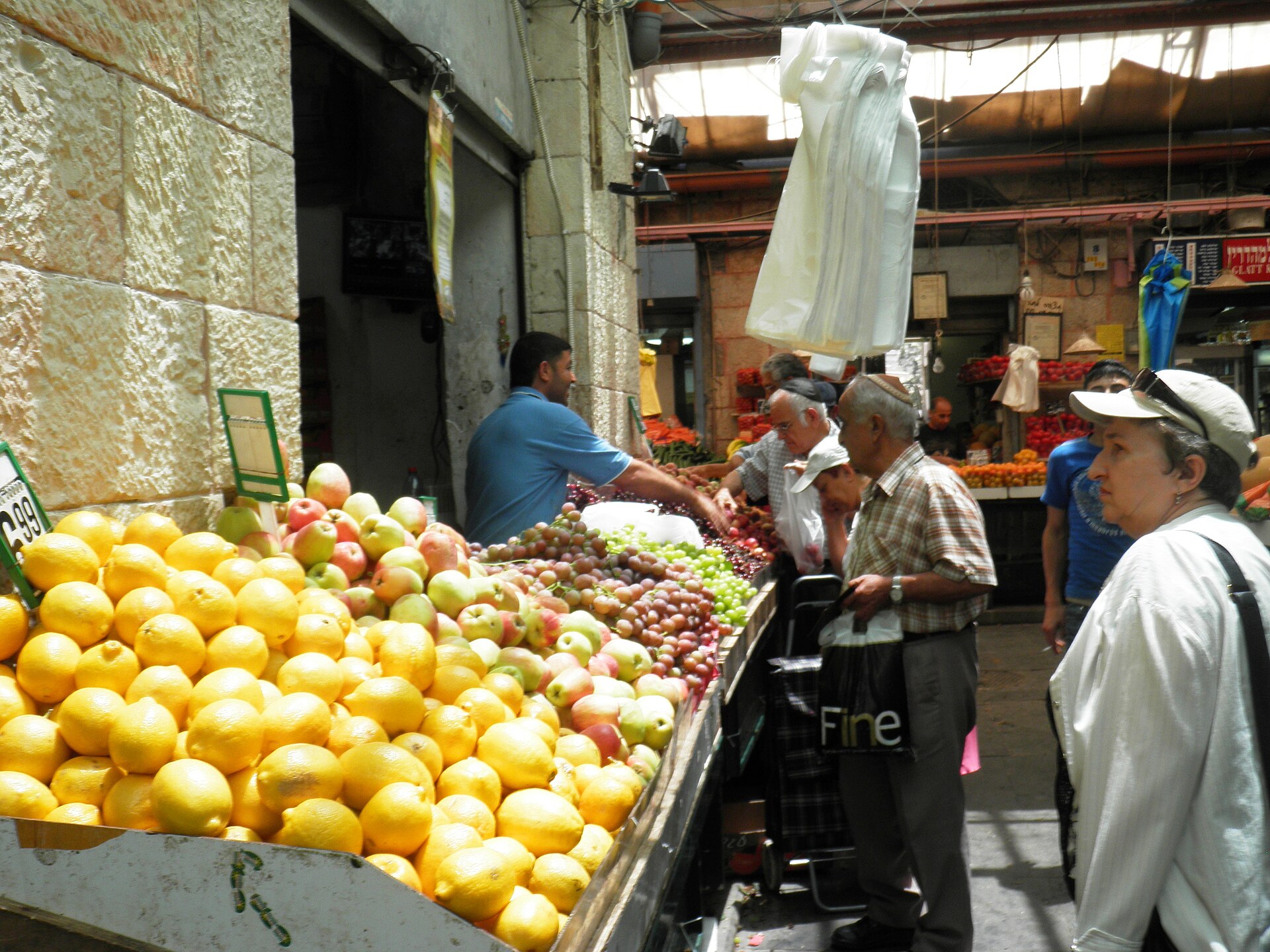Shoppers and a vendor at a Mahane Yehuda Market fruit stall piled with lemons, apples and grapes, in a covered lane with Jerusalem stone walls