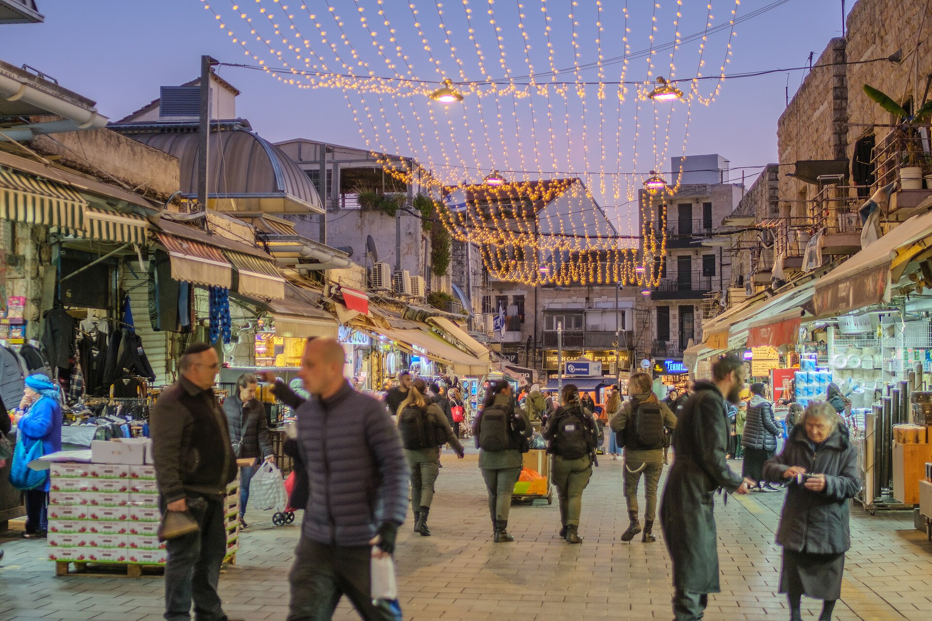 Shoppers walk along Mahane Yehuda Street at dusk beneath canopies of fairy lights, with lit clothing and household stalls on both sides and a domed market roof