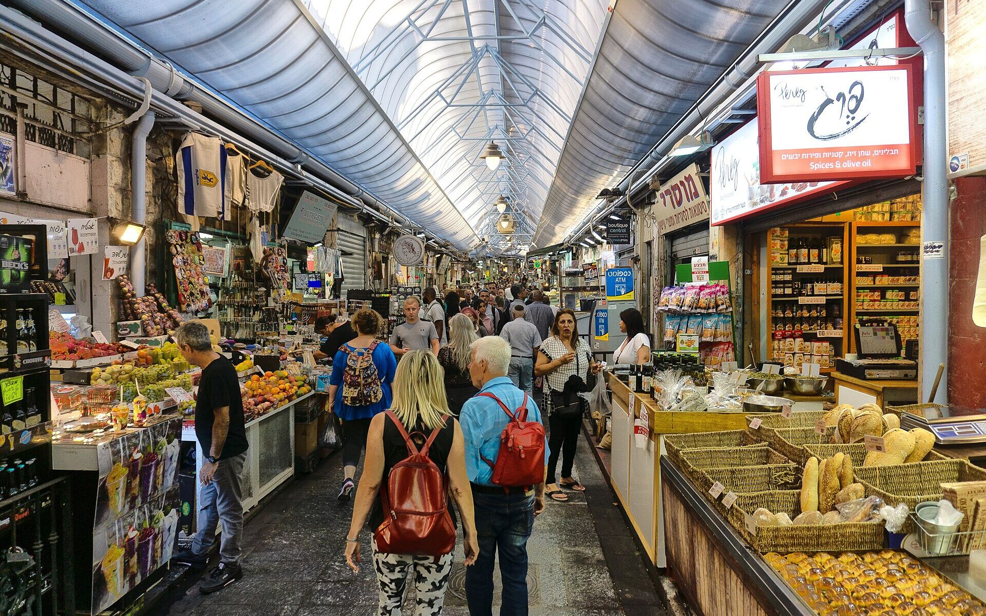 Shoppers walk through a covered lane of Mahane Yehuda Market in Jerusalem, past fruit stalls, baskets of bread and a spice and olive oil shop with Hebrew signs