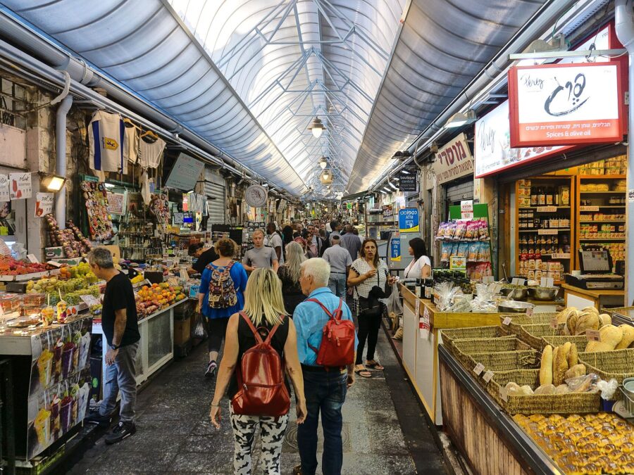 Shoppers walk through a covered lane of Mahane Yehuda Market in Jerusalem, past fruit stalls, baskets of bread and a spice and olive oil shop with Hebrew signs