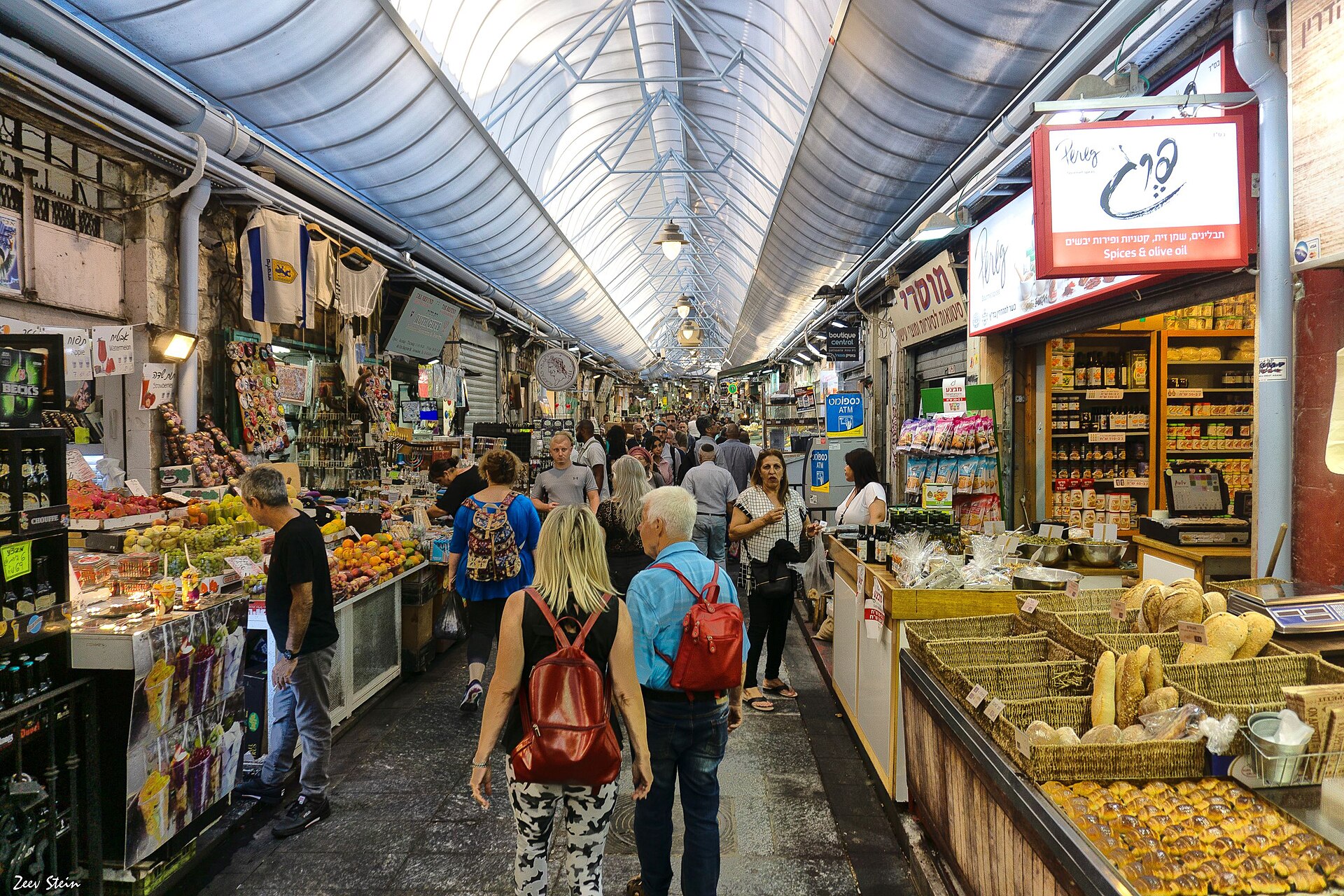Shoppers walk through a covered lane of Mahane Yehuda Market in Jerusalem, past fruit stalls, baskets of bread and a spice and olive oil shop with Hebrew signs