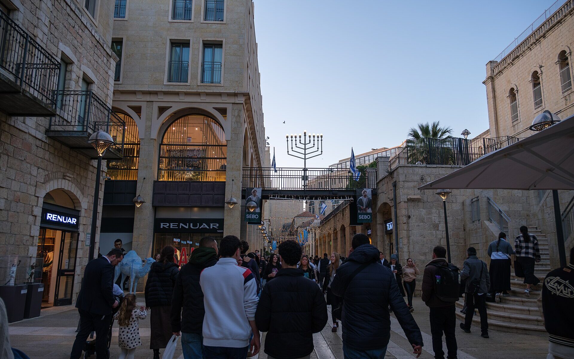 Shoppers walking along Mamilla Avenue in Jerusalem between stone buildings with shopfronts, a blue camel sculpture and a footbridge topped by a large Hanukkah menorah