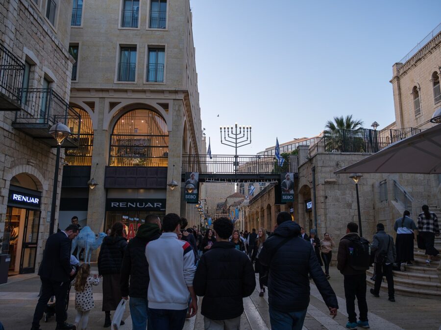 Shoppers walking along Mamilla Avenue in Jerusalem between stone buildings with shopfronts, a blue camel sculpture and a footbridge topped by a large Hanukkah menorah
