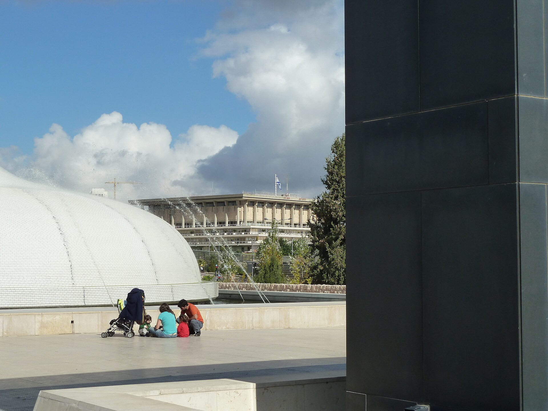 The white dome of the Shrine of the Book at the Israel Museum with fountains playing, a family sitting on the plaza, and the Knesset building on the hill behind