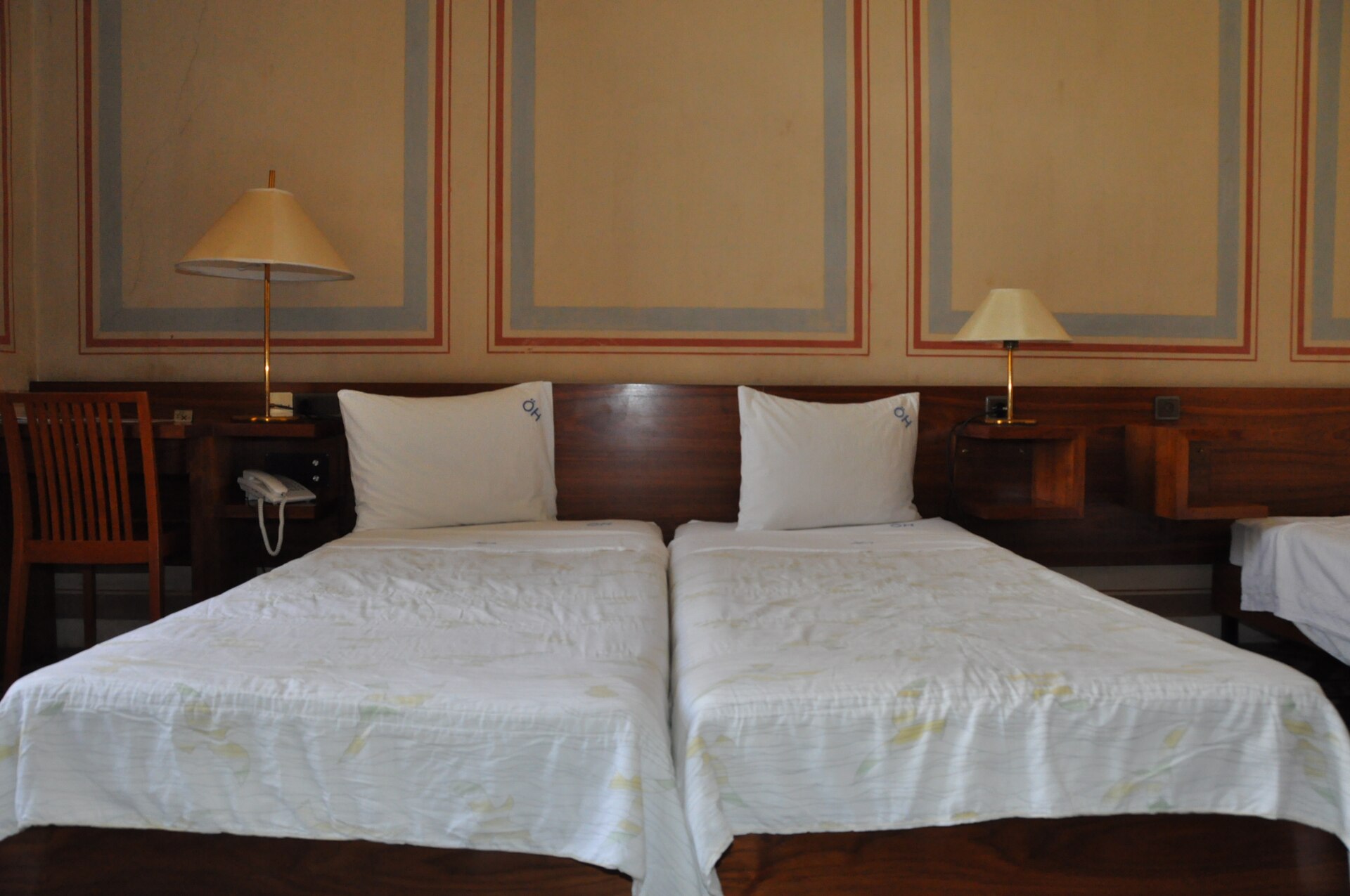 Simple twin guest room at the Austrian Hospice in Jerusalem, with two single beds, white covers, pillows monogrammed ÖH, bedside lamps and painted panels on the wall
