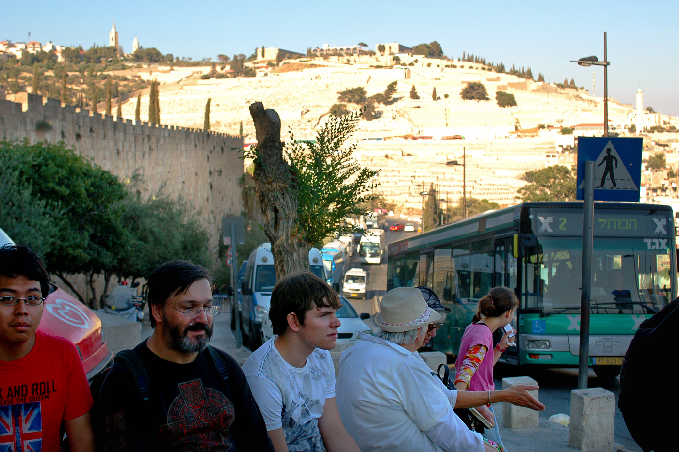 A small group of visitors resting on a low wall outside Jerusalem's Old City walls, with the Mount of Olives and its cemeteries rising behind and a city bus passing