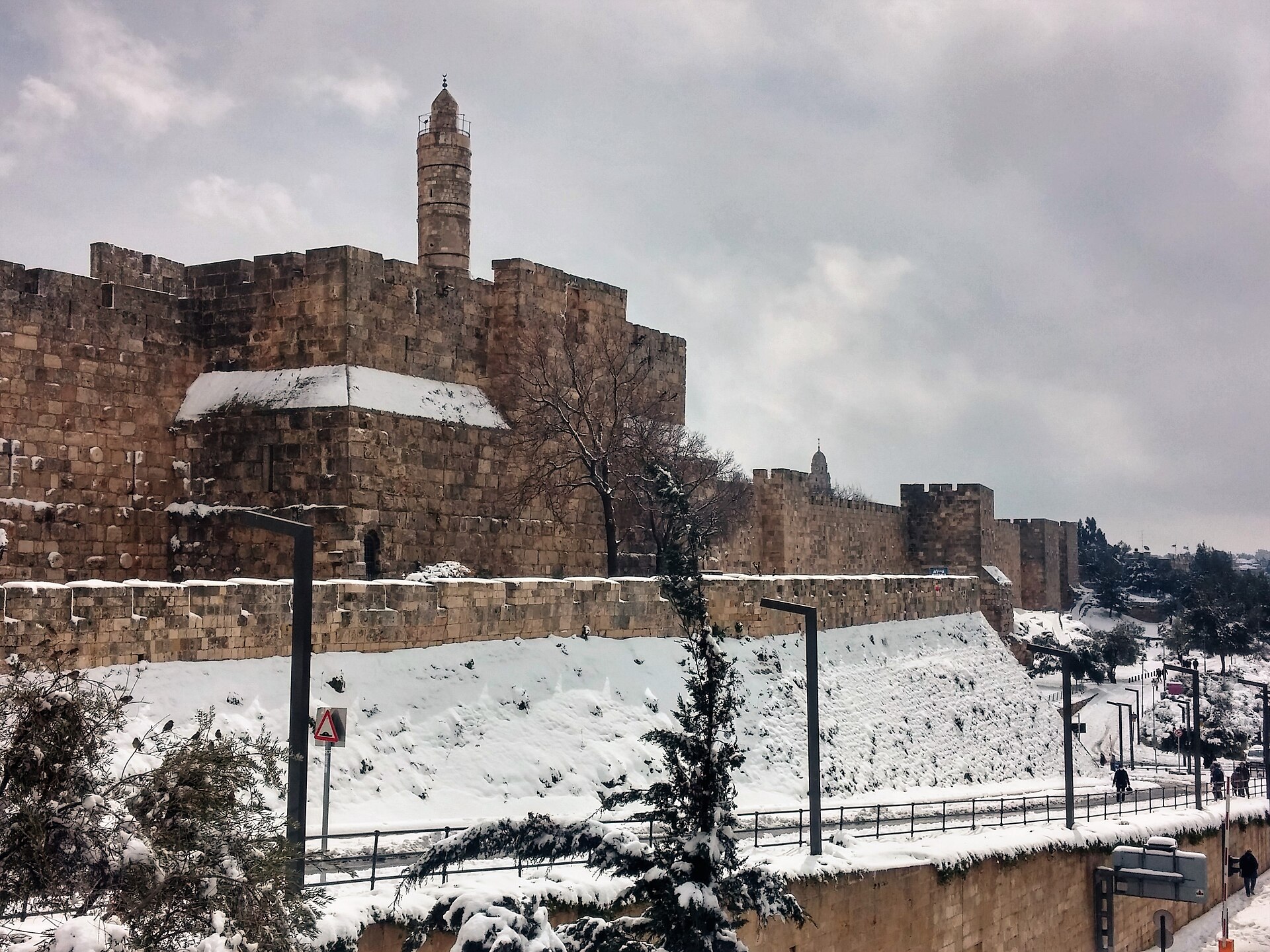 Snow covers the sloping glacis below the Tower of David citadel and Jerusalem's Old City walls under a grey February sky, with snow-laden trees in front