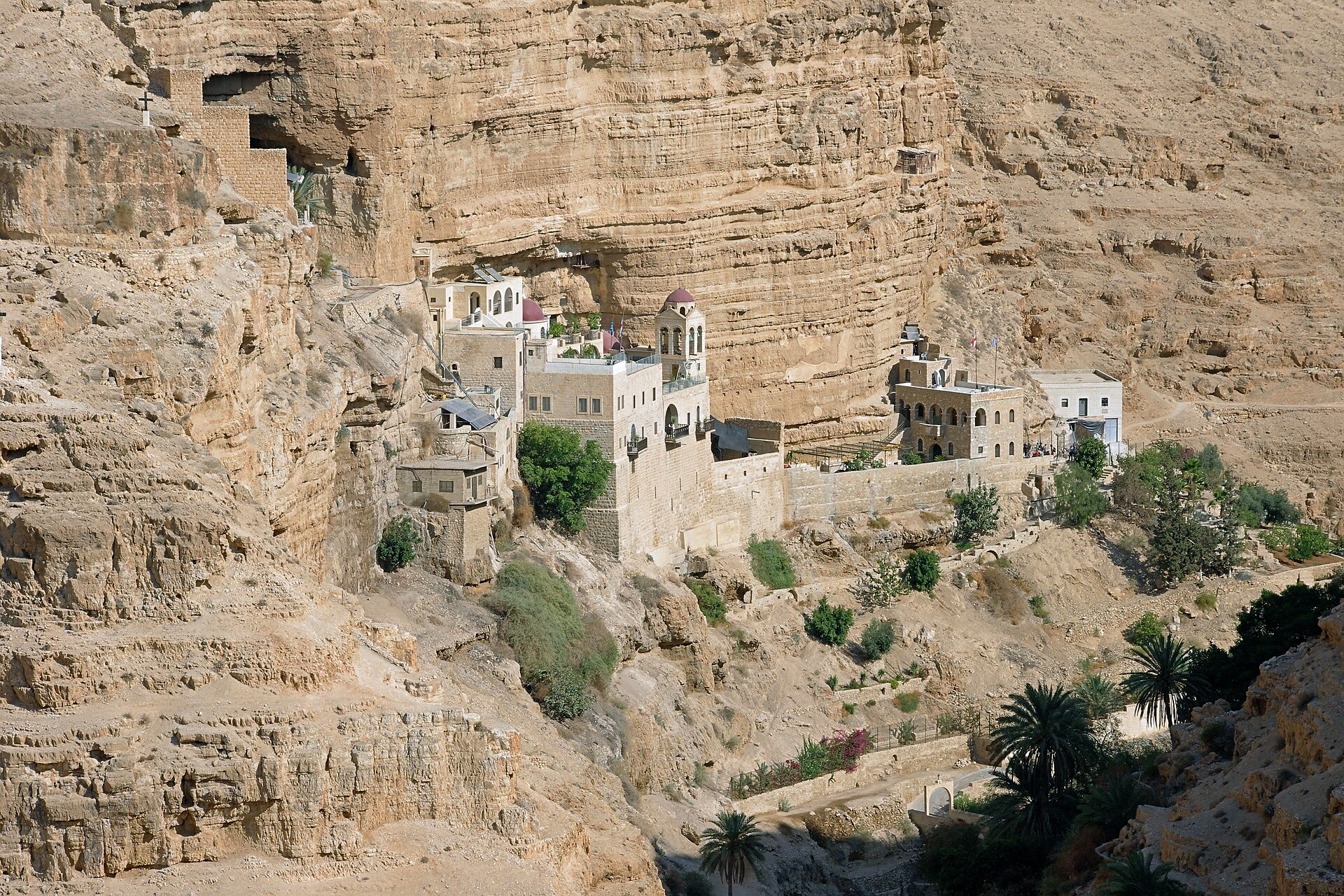 St George's Monastery built into a sheer limestone cliff in Wadi Qelt, with red domes, a bell tower, a cave chapel above and palm trees below