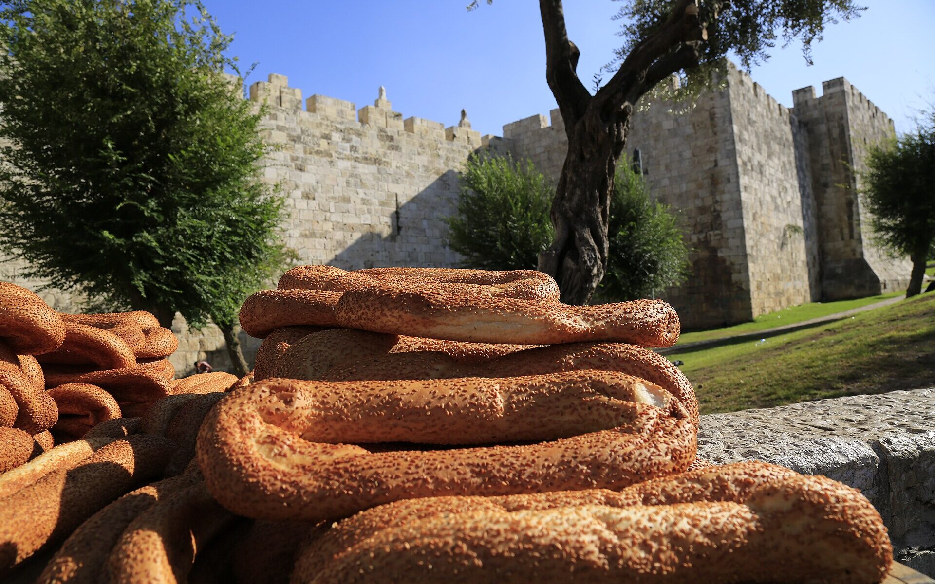 Stacks of sesame-crusted ka'ak bread in the foreground, with the crenellated stone walls of Jerusalem's Old City and an olive tree behind