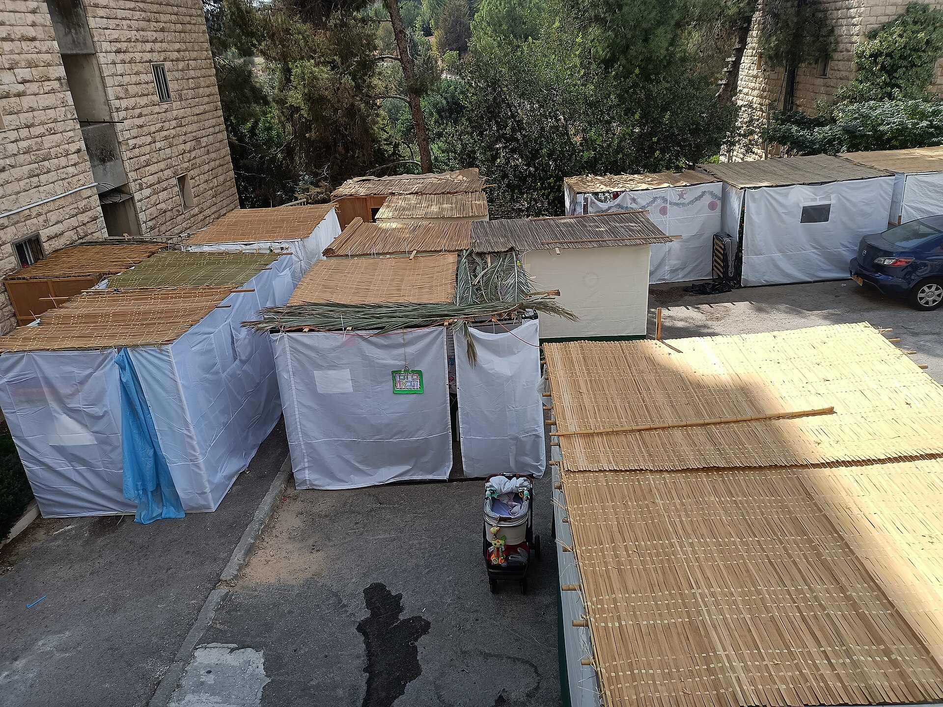 Sukkah booths with bamboo mat and palm frond roofs and white sheeting walls filling a car park beside Jerusalem apartment buildings