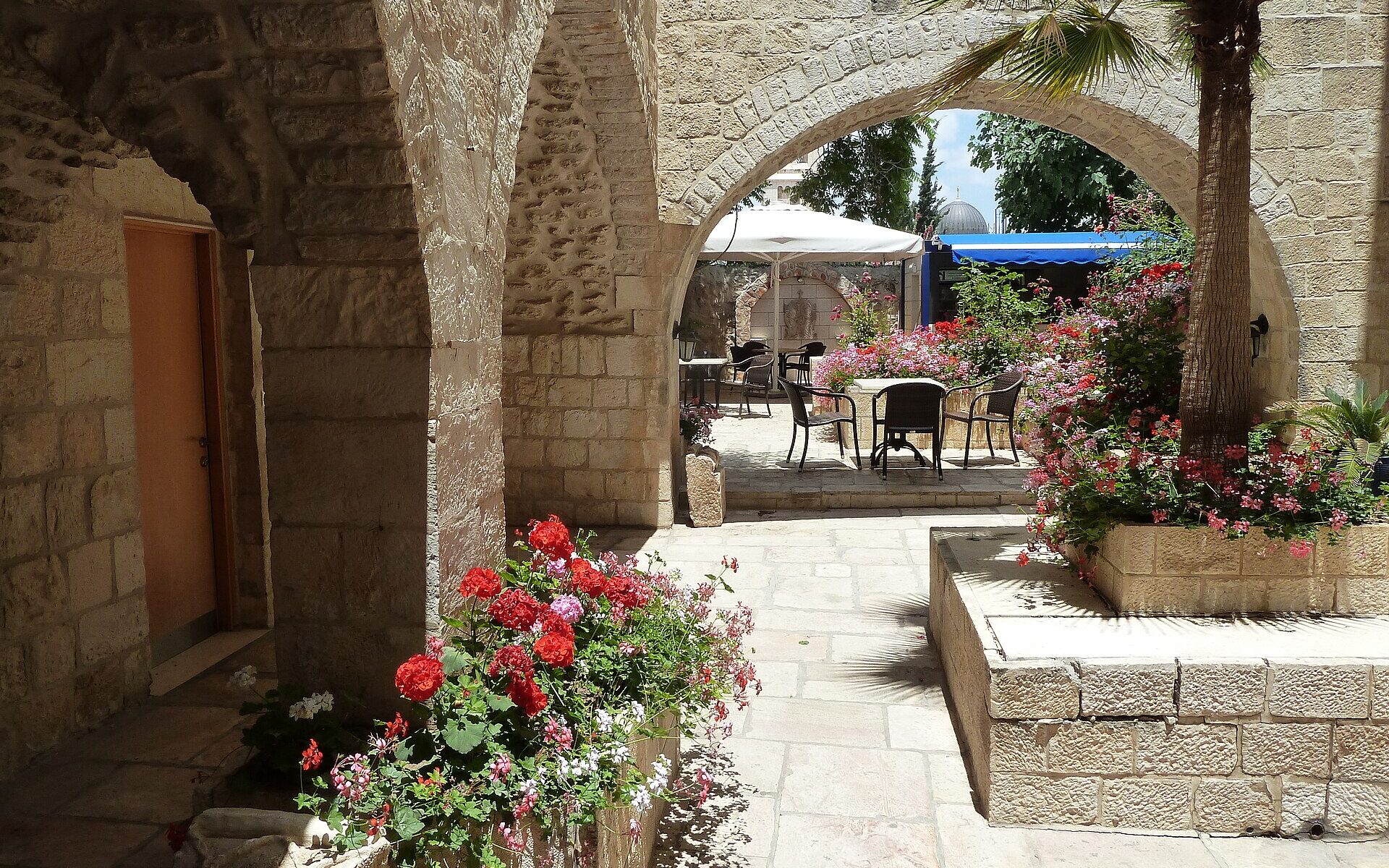Sunlit stone courtyard of the Lutheran Guesthouse in Jerusalem's Old City, with arches, a palm in a raised planter, red geraniums and café tables beyond