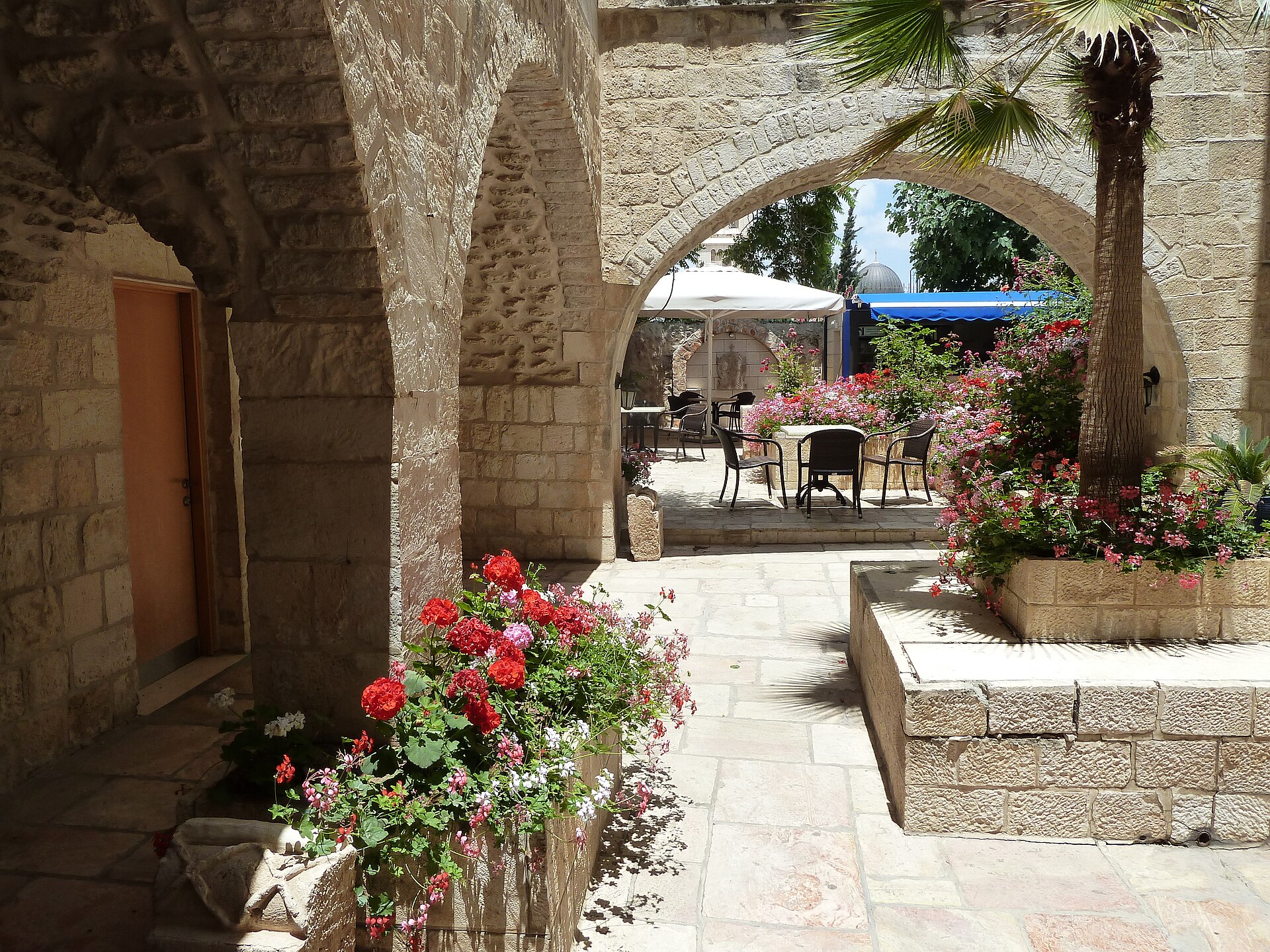 Sunlit stone courtyard of the Lutheran Guesthouse in Jerusalem's Old City, with arches, a palm in a raised planter, red geraniums and café tables beyond