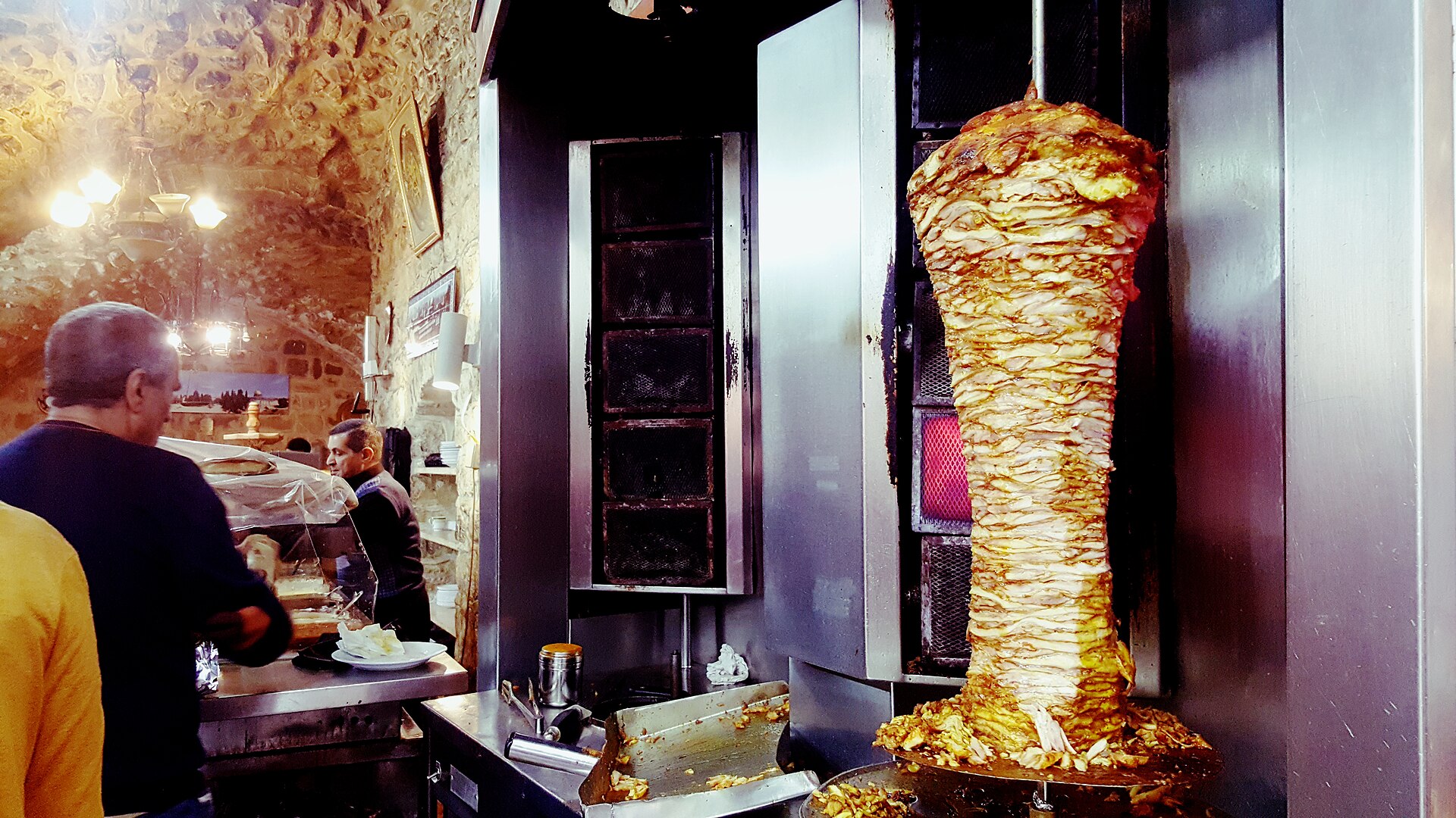 Tall shawarma spit roasting beside a vertical grill inside a stone-vaulted Old City shop, with a server at the counter and customers waiting