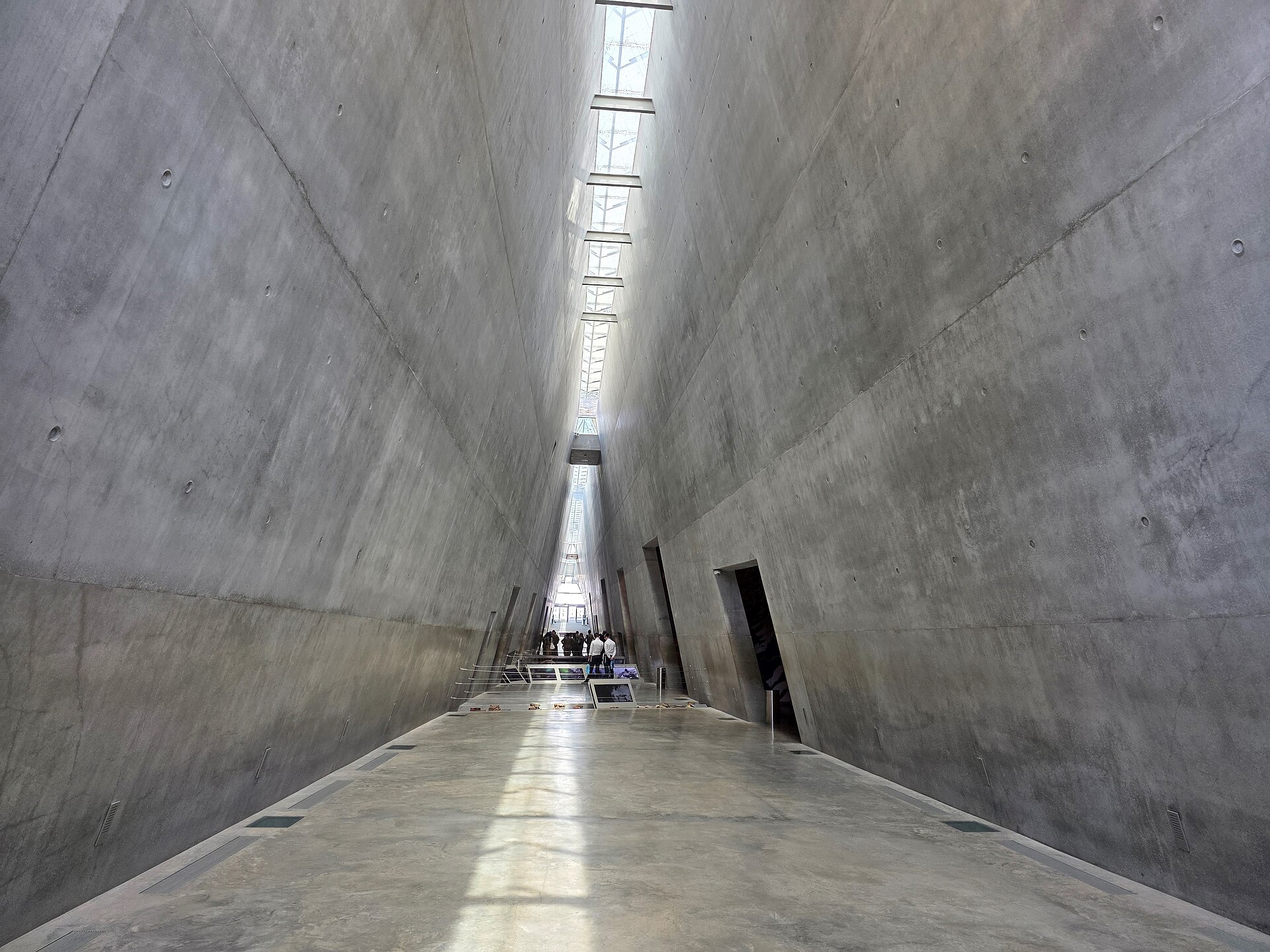 Tall triangular concrete corridor of the Holocaust History Museum at Yad Vashem, lit by a narrow skylight along its apex, with gallery openings leading off to the side