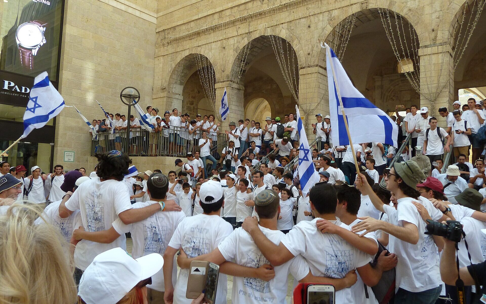 Teenagers in white T-shirts dance arm in arm and wave Israeli flags beneath the stone arcades of Mamilla, just outside the Old City, on Jerusalem Day
